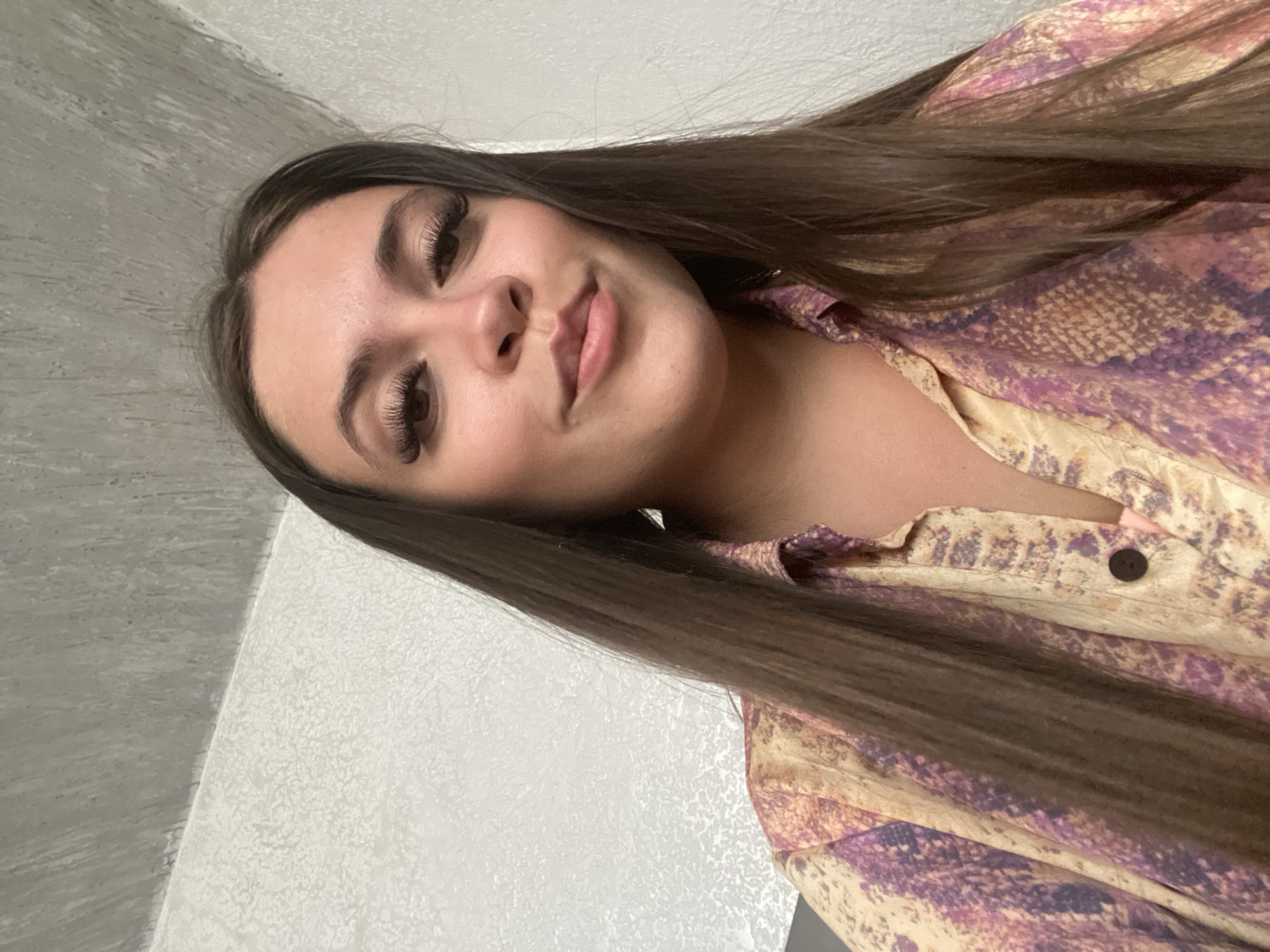 A medium shot captures a young woman from the shoulders up, looking towards the camera. Her long, straight, dark brown hair frames her face and falls down her shoulders. She has prominent eyelashes, dark eyebrows, and is wearing a button-up shirt with a colorful, textured pattern of pink, purple, and tan. The background is a textured white wall, with a portion of a gray, textured ceiling visible in the upper left quadrant. The scene appears to be indoors, in a brightly lit room. The specific location in the city of Chalda, Russia cannot be determined from the visual information provided.