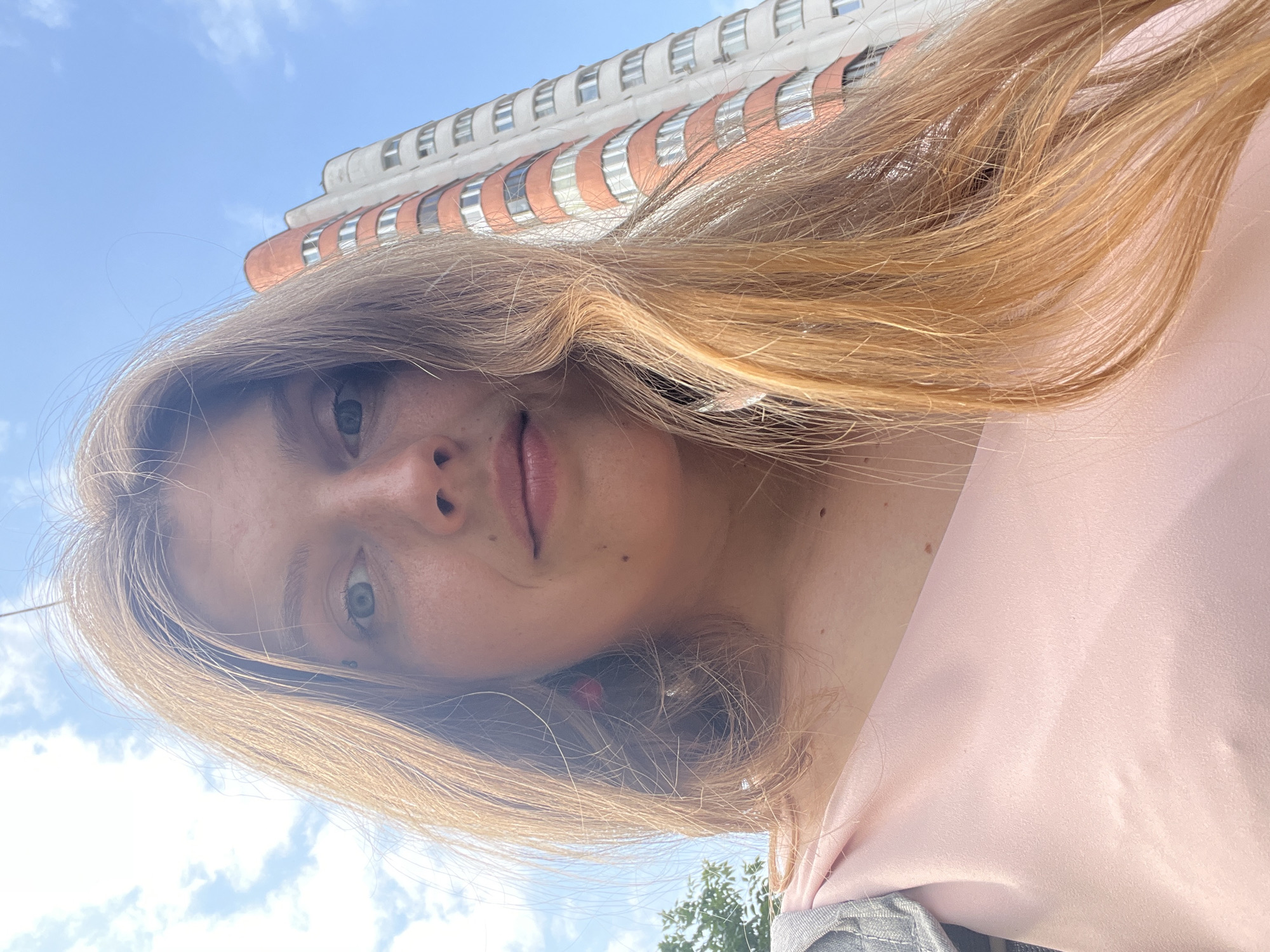 A close-up, low-angle, selfie-style photograph captures a young woman with long, auburn hair against a bright blue sky with scattered clouds. She is looking directly at the camera. A tall, multi-story apartment building with balconies and windows is visible in the background, to her right. The building features a mix of white and orange facades. A hint of green foliage is visible to the far left. The woman is wearing a pale pink top and a grey garment, possibly a jacket or cardigan, is draped over her left shoulder. A small, bright pink earring is visible on her left ear. The scene appears to be outdoors in an urban environment, consistent with a street or residential area in Minsk, Belarus.