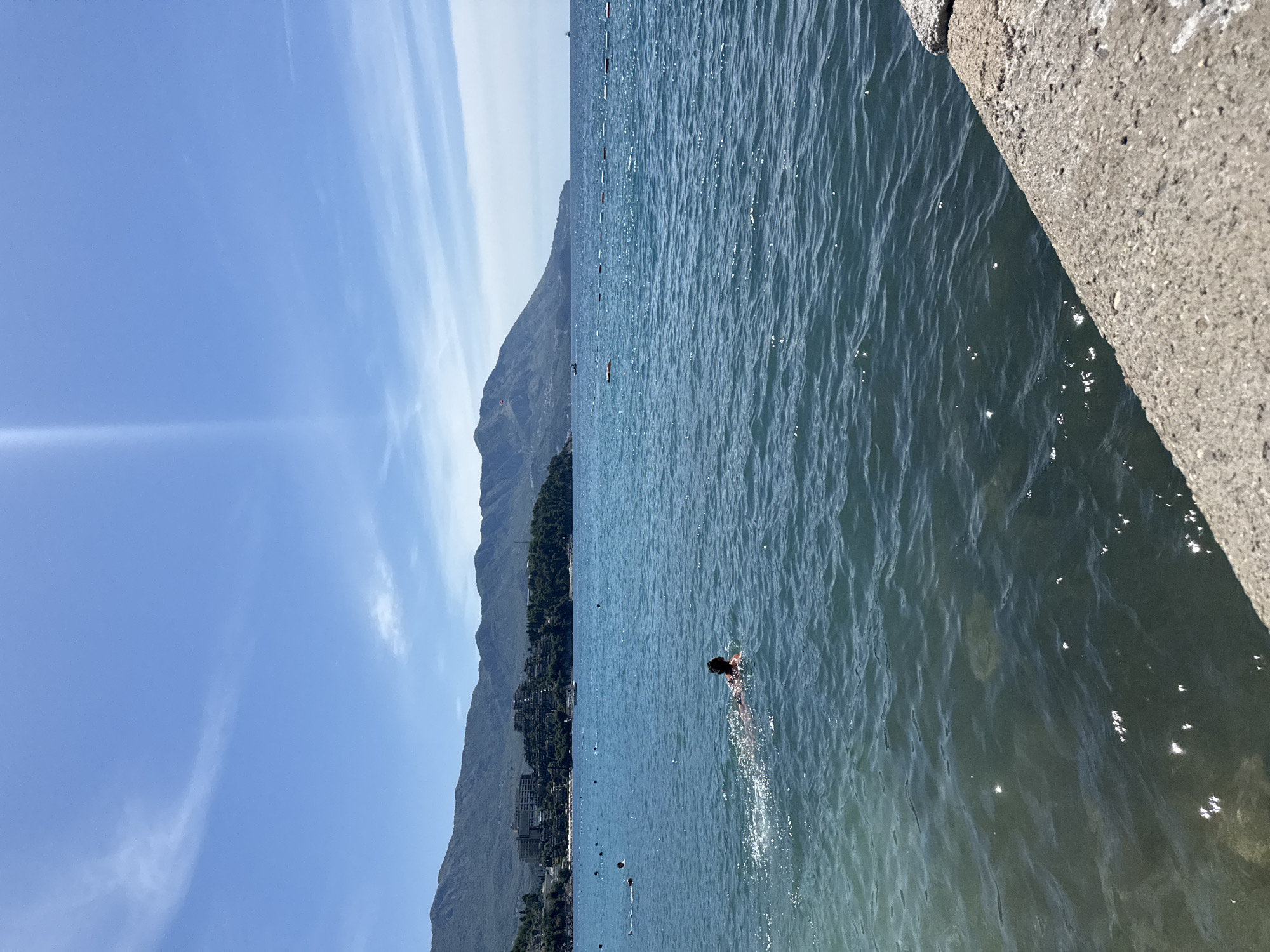 In Budva, Montenegro, a person wearing a snorkeling mask swims in the clear blue waters of the Adriatic Sea. The person, seen from behind, is on their stomach, propelling themselves forward. Further out in the water, other swimmers and buoys are visible, indicating a popular beach area. In the background, lush green hills rise up, dotted with hotels and buildings, suggesting a developed coastal resort. The sky above is a bright, clear blue with wispy white clouds. The foreground shows a stone embankment, likely a pier or seawall, where the photo was taken from.