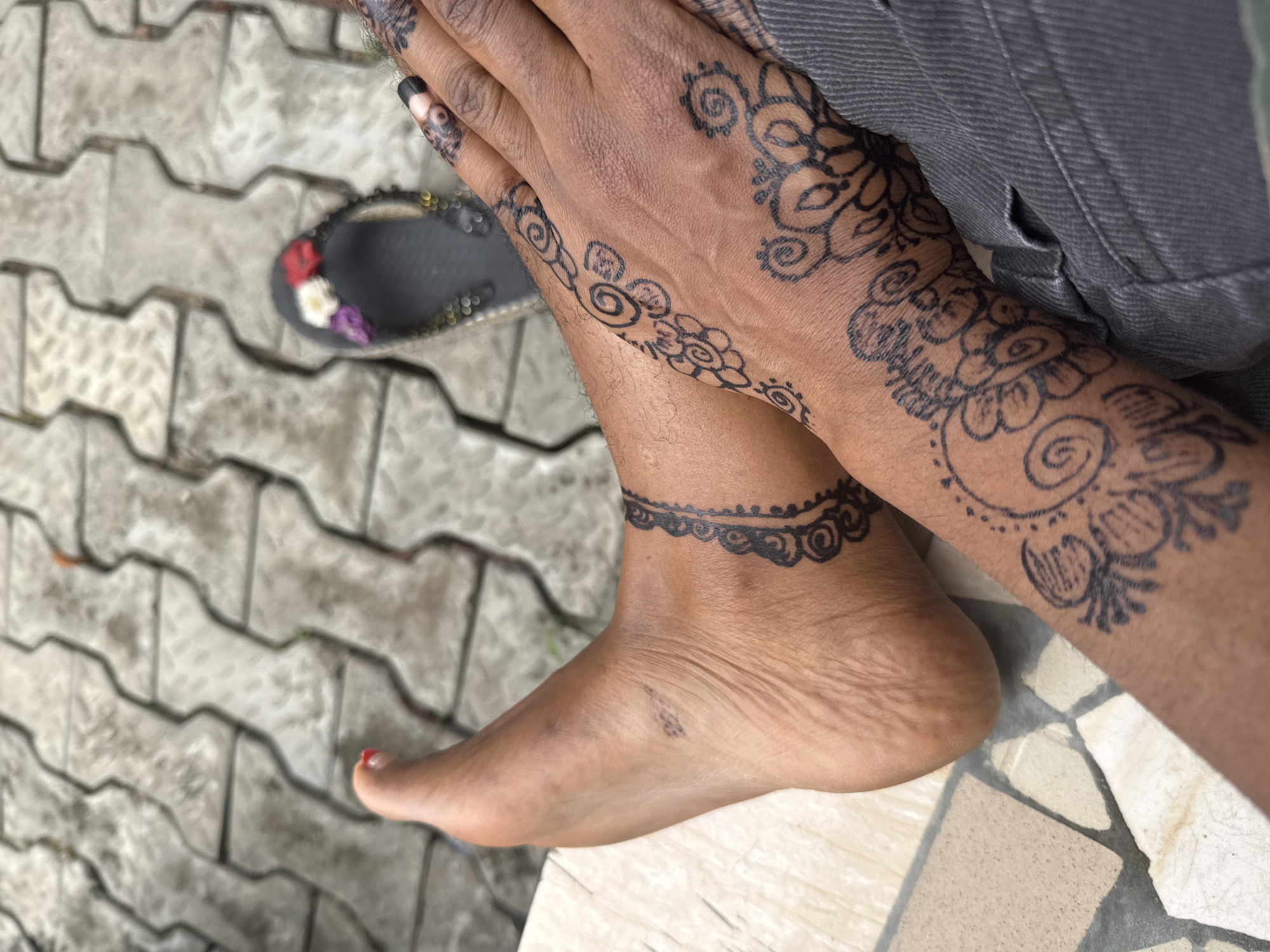 This close-up, high-angle shot captures the lower body of a person, likely in Ado, Nigeria, adorned with intricate black henna (mehndi) designs. The subject's hands, lower arms, and one ankle are prominently featured, all decorated with traditional, swirling patterns. The henna extends across the backs of the hands, fingers (with black nail polish visible on the fingernails), and forms an anklet-like band around the right ankle. A single toenail on the right foot shows a small spot of red nail polish.
The person is wearing a gray fabric garment, possibly trousers or a skirt, which covers their upper legs. A black flat sandal or shoe, embellished with small red, white, and purple floral decorations, rests on the ground beside the left foot.
The setting appears to be an outdoor paved area, with a distinctive pattern of light gray interlocking concrete blocks visible in the foreground and background. A lighter-colored, possibly tiled or stone surface, is also beneath the foot and ankle in the bottom right corner of the frame. The lighting suggests it is daytime, though the exact time is unclear. No text is visible in the image.