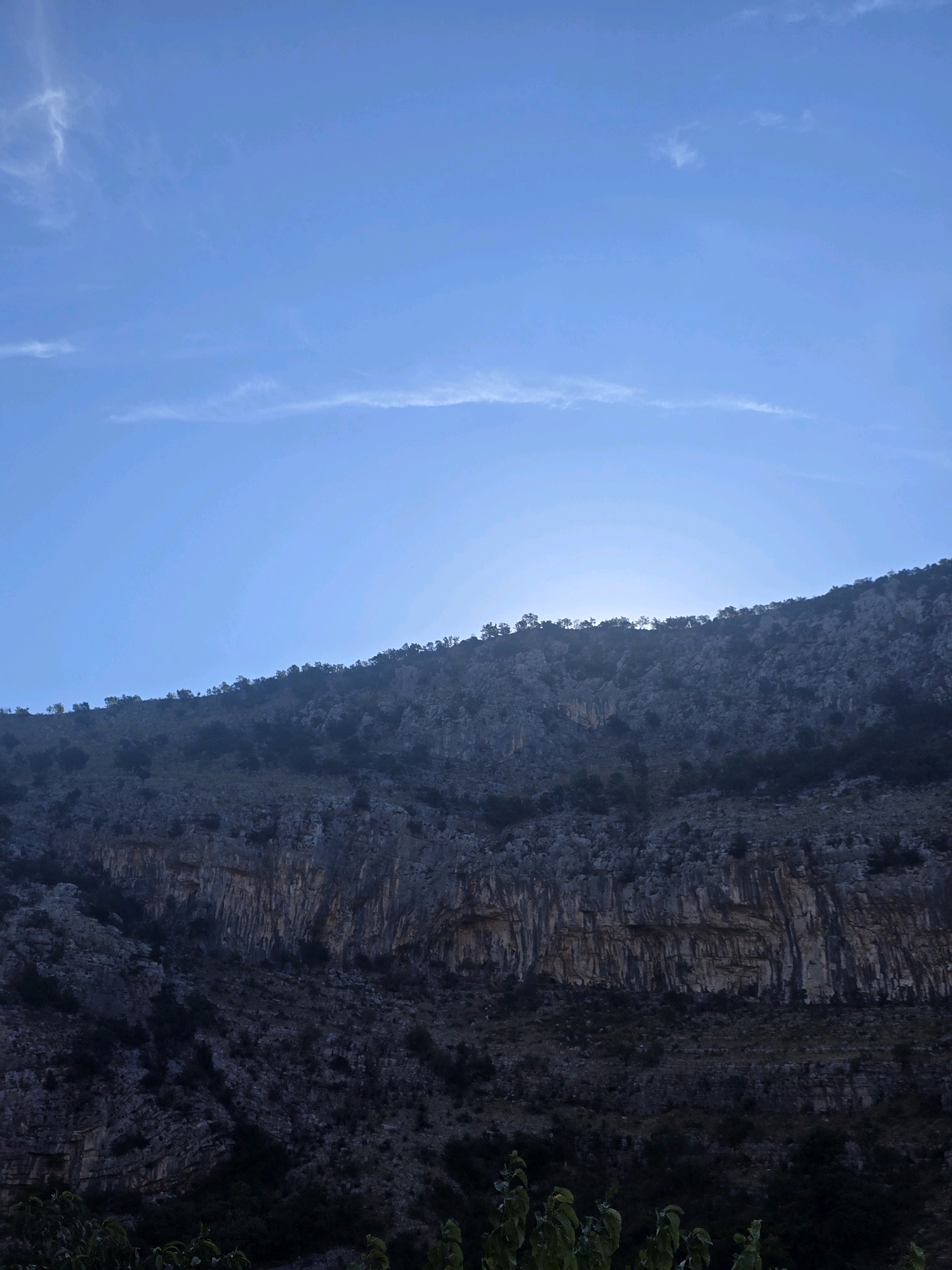 The image shows a rocky, mountainous landscape under a clear blue sky with faint wispy clouds. The sky occupies the upper half of the image, gradually transitioning to a slightly brighter hue near the horizon where it meets the mountains. The mountains themselves are steep and rugged, with visible rock formations and vegetation growing on their slopes. The lower portion of the mountains is darker, suggesting shadows or a denser collection of trees. In the foreground, a few green leaves are visible, partially obscuring the bottom edge of the frame. The scene appears to be a natural landscape, likely in a mountainous region, with no signs of urban development or human presence. The location is identified as the city of Podgorica, Montenegro.