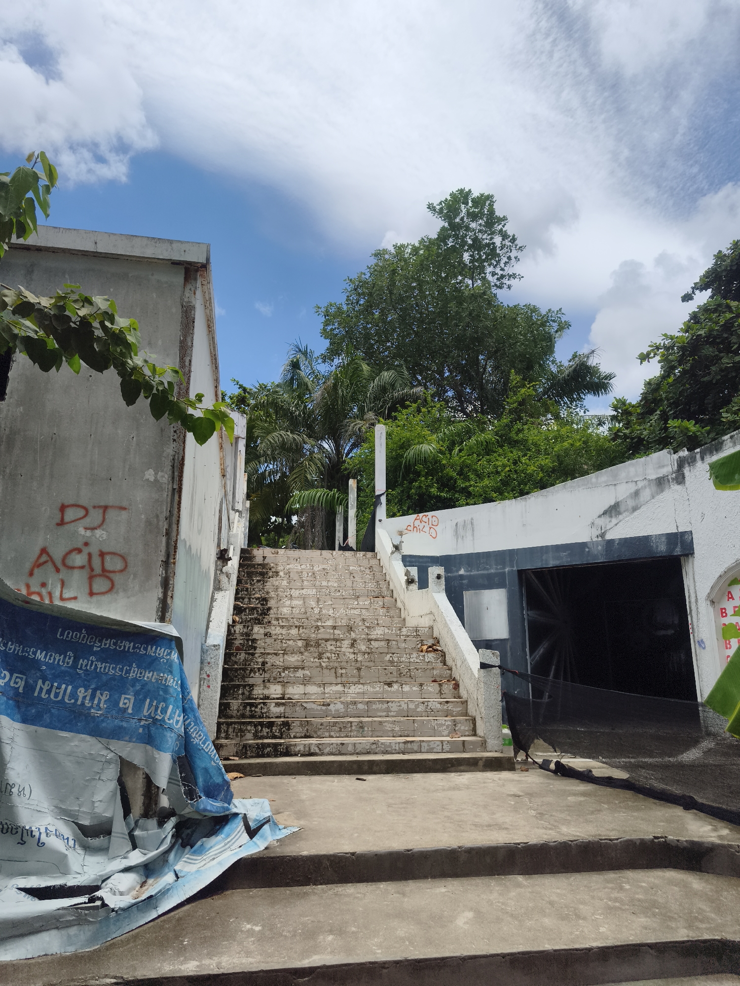 The image shows an outdoor staircase in a tropical environment, likely on the island of Ko Samui, Thailand, as indicated by the lush vegetation and weathered concrete structures. The staircase, made of concrete, ascends to the right, flanked by white walls. Graffiti, including red spray paint reading "DJ ACID CHILD," is visible on the left wall and on a white structure to the right. A large, torn blue and white tarp is draped over the lower part of the left wall and extends onto the ground. The stairs themselves appear old and worn, with some debris scattered on them. The environment is characterized by green trees and a bright blue sky with scattered white clouds. To the right of the staircase, there is a dark opening, possibly an entrance, covered by a black net. The overall scene suggests a somewhat neglected or abandoned outdoor space in a warm climate.