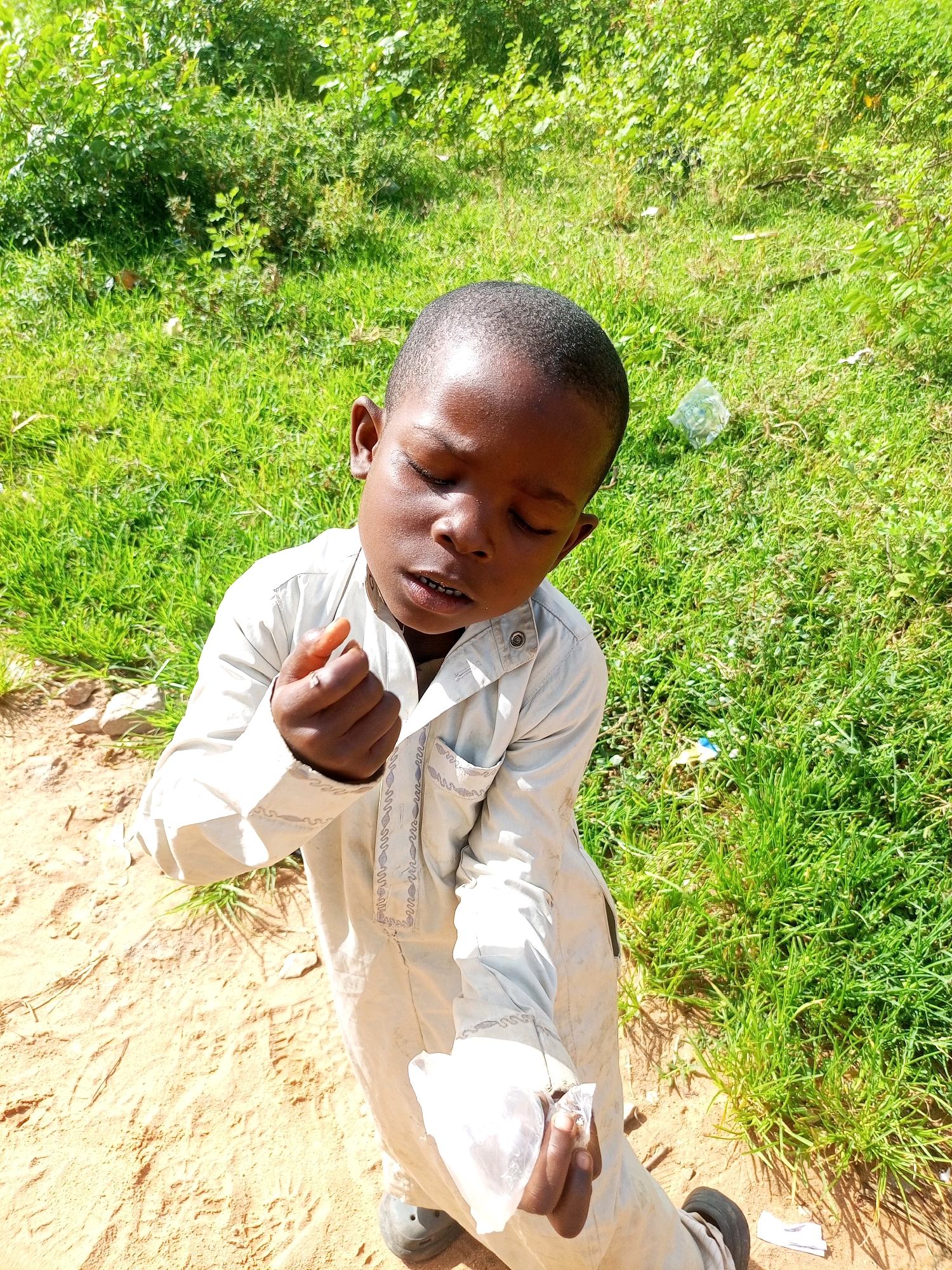A young Black child, appearing to be between 5 and 7 years old, stands on a dirt path in a grassy, outdoor environment. The child is wearing a light-colored, long-sleeved garment with decorative stitching around the neckline and pocket. The child's eyes are closed, and their head is tilted downwards, with their right hand raised, holding a small object between their thumb and index finger. Their left hand is lowered, also holding something wrapped in white material. The ground beneath the child consists of sandy, light brown dirt with scattered rocks and some sparse green vegetation. To the right and behind the child, there is lush green grass and dense green foliage, suggesting a natural or semi-rural setting, possibly on the outskirts of Tempure, Nigeria. The lighting indicates it is daytime, with sunlight casting shadows. Scattered in the grass are a few pieces of white plastic debris. The overall impression is one of a candid moment captured outdoors.