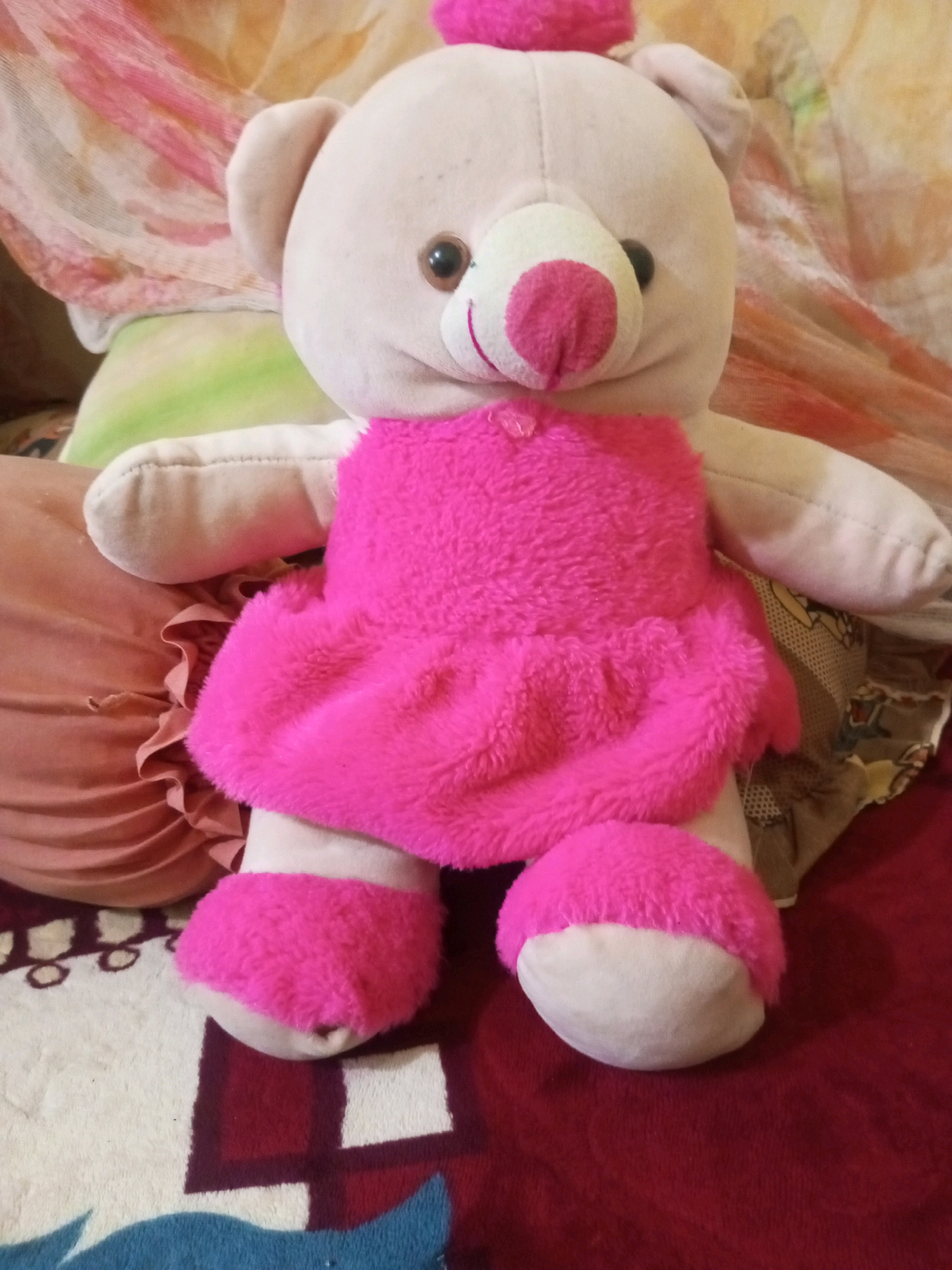 A light pink stuffed teddy bear with large, dark eyes and a pink nose is seated on a red blanket. The bear is wearing a bright pink, fuzzy dress and matching fuzzy foot coverings. The background is out of focus, but appears to be a mix of patterned fabrics and pillows in muted colors. The scene is indoors, possibly in a bedroom or living area. There are no people visible in the image. The location in the city of Bundar, Indonesia is not discernible from the image.