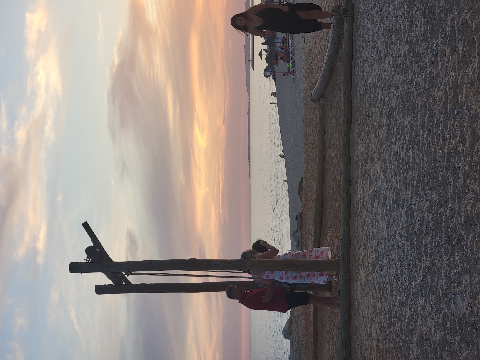 An outdoor, eye-level, full shot shows a beach scene at dusk in Quarteira, Portugal. The sandy beach in the foreground is marked by a horizontal wooden log. In the midground, a wooden structure resembling a swing set with two vertical posts and a horizontal crossbeam is visible. Three people are standing near the wooden structure. A man in a red polo shirt and dark shorts stands to the left, facing a woman in a white and pink floral dress. A third person, partially obscured, is also near them. Further back on the beach, other people are scattered, some sitting under umbrellas. The calm sea stretches to the horizon under a sky filled with soft, pastel-colored clouds. A woman in a long black dress walks towards the right side of the frame.