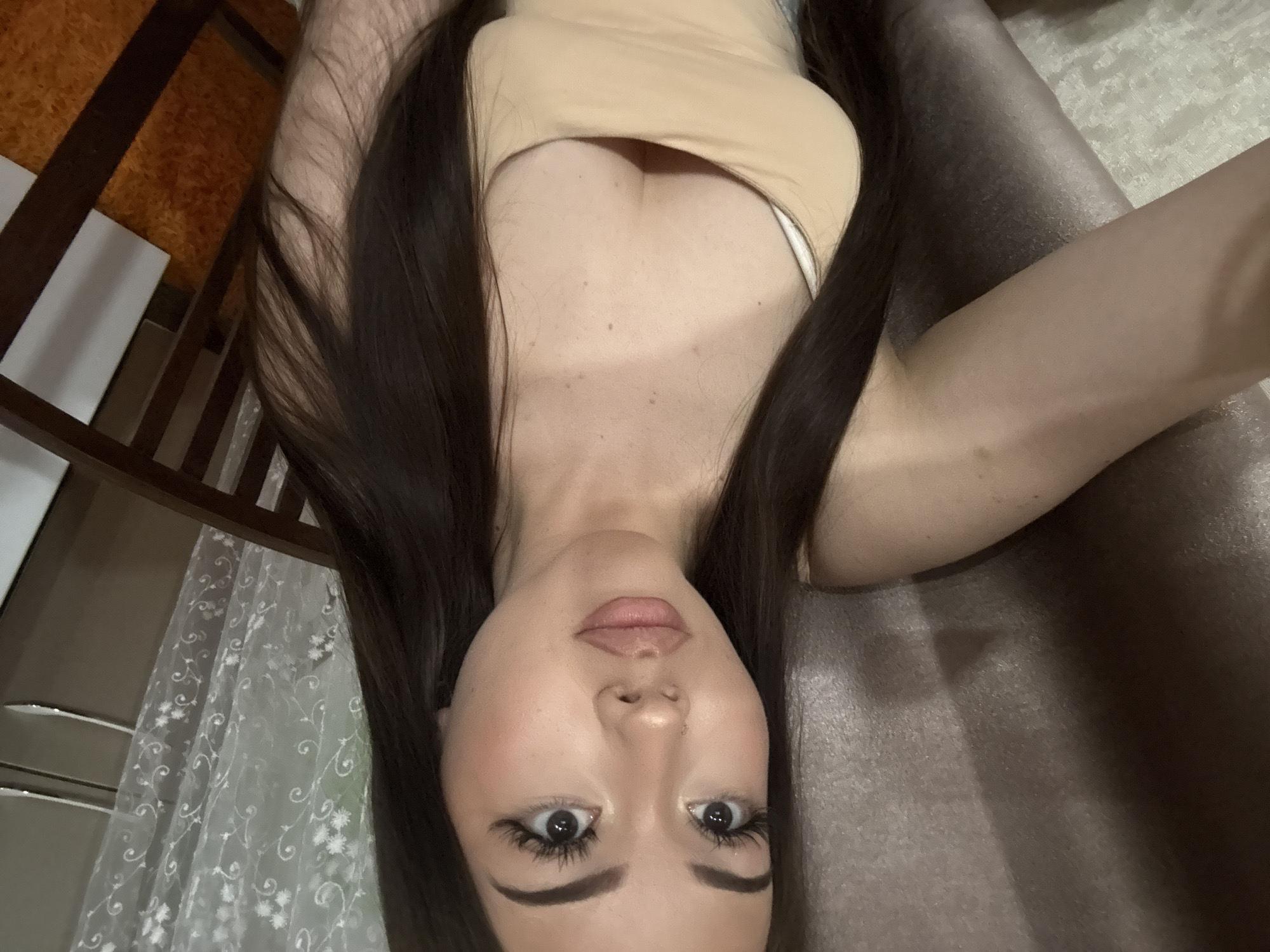 A young woman is depicted in a close-up, top-down selfie, lying down in an indoor setting. Her head is at the bottom of the frame, and she gazes directly at the viewer with wide, dark eyes. She has long, dark brown hair flowing around her, and her face is adorned with dark, defined eyebrows, extended eyeliner, and full, slightly parted lips with a natural-toned lip gloss. She wears a light beige or nude-colored tank top, revealing visible cleavage and a white strap underneath. Her skin appears fair with a few faint freckles or moles on her chest. The background features a dark brown, possibly leather or fabric, surface to her right, suggesting a couch or bed. To her left, a dark wooden structure, possibly a bed frame, is visible, partially obscuring a sheer white curtain with a delicate floral pattern. An orange-brown textured wall is faintly seen in the upper left background. The overall scene conveys a relaxed pose in a home environment, likely in Naberezhnye Chelny, Russia, during daytime hours. There is no visible text.