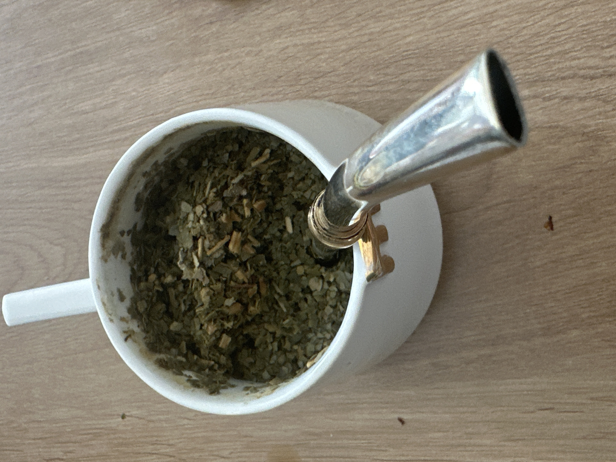 The image captures a top-down view of a white ceramic cup containing dried yerba mate leaves, with a silver-colored metal bombilla (straw) inserted into the mixture. The cup is positioned on a light-colored wooden surface, likely a table or countertop.
The cup is simple and white, with a handle partially visible on its left side. Inside, the loose, green and brown yerba mate appears dry, suggesting it is either awaiting its first infusion of hot water or has been prepared and is ready for consumption. The bombilla, a distinctive feature of mate preparation, is metallic and reflective, angled upwards and towards the right side of the frame. Its wide, flattened mouth is visible at the top. The portion of the bombilla submerged in the mate indicates the presence of a filter.
The wooden surface underneath has a light, natural grain pattern, with slight variations in tone. There are no other objects or people visible in the frame. The lighting is diffused and even, with soft reflections on the bombilla. The scene is static, depicting a moment before or during the consumption of yerba mate. No text is visible in the image. This scene takes place in Fethiye, Turkey.