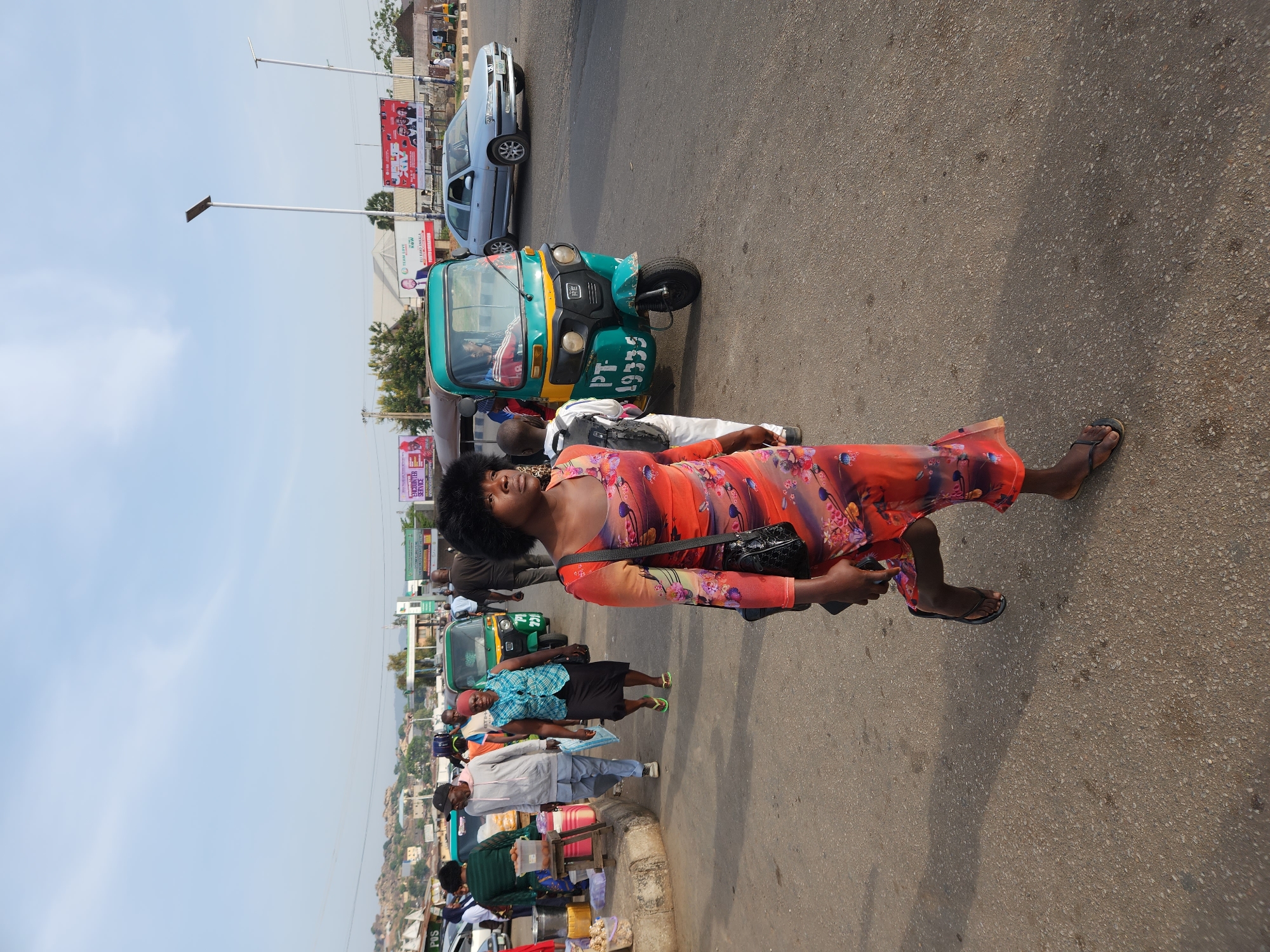 Captured in the city of Jos, Nigeria, the image presents an outdoor scene on an asphalt road beside a sidewalk. A woman wearing a floral patterned dress and sandals is walking on the road. Nearby is a green three-wheeled vehicle and a silver car. The sidewalk is lined with vendors and pedestrians. Above this scene, billboards and utility poles punctuate the skyline.