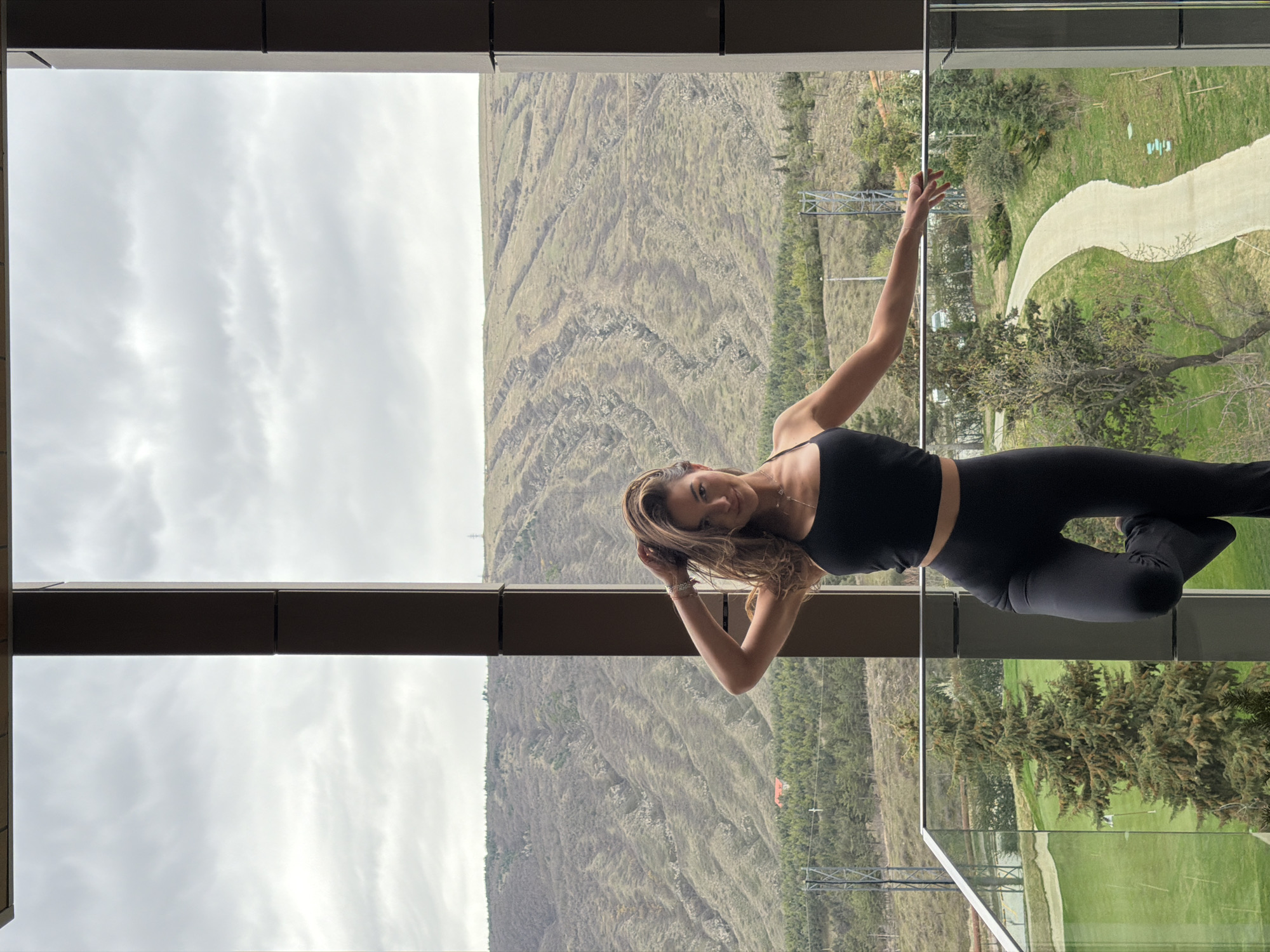 A female individual is positioned centrally within a large window or balcony frame, facing forward with a slight smile. Her long, wavy brown hair is lifted by her right hand, which is raised to the back of her head. She is wearing a black spaghetti-strap crop top and black flared leggings. Her left leg is bent at the knee, with the foot resting against her right inner thigh, while her left arm is extended, resting on the silver-colored horizontal railing of a glass balcony.
The background presents a panoramic view of the landscape consistent with the Tbilisi Hills Golf & Residences area in Tbilisi, Georgia. Immediately below the balcony, a manicured green lawn, characteristic of a golf course, is visible, interspersed with evergreen trees. A light-colored, winding pathway extends through a grassy area in the lower right. Beyond this, a series of barren to sparsely vegetated hills and mountains rise, displaying visible erosion patterns and scattered clusters of trees. A red-roofed building is discernible on a distant lower slope to the left, and metal lattice-work structures (pylons) are visible on the far slopes. The sky is uniformly overcast with grey clouds.