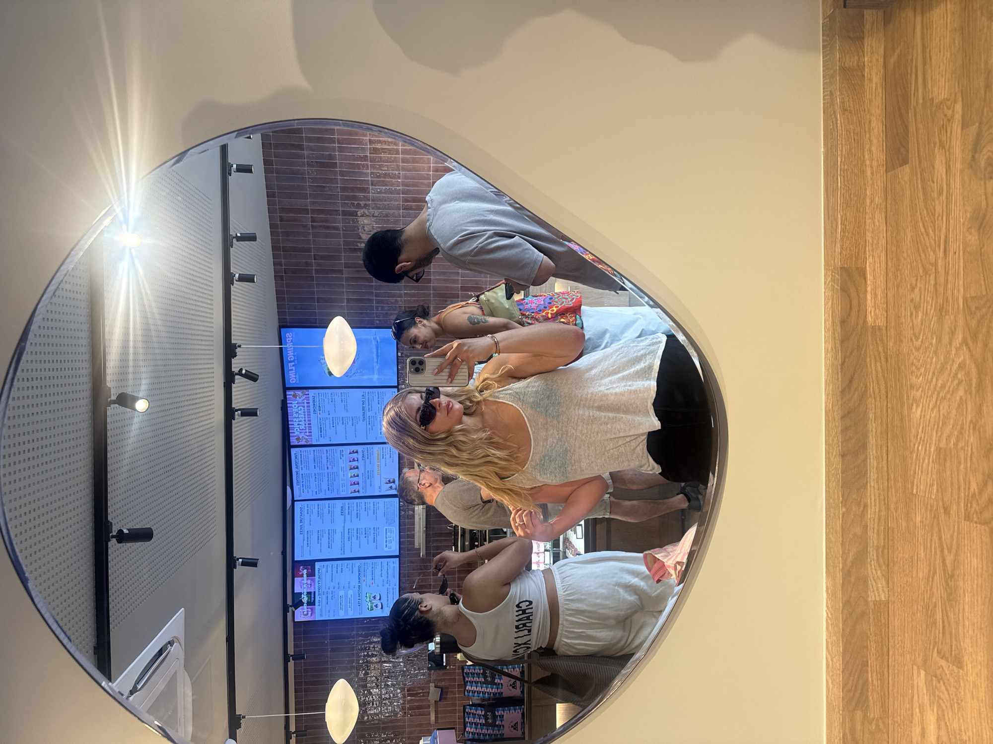 Here is a detailed description of the image:
**Location:** The image appears to be taken inside a restaurant or café, likely in Paris, France, based on the text in the mirror which has what appears to be reversed French phrases. The interior features what appears to be menu boards and brick-like tiles.
**People:** The image features several people reflected in an oval mirror.
* There is a man with short hair wearing a light-colored t-shirt.
* A woman with dark hair pulled back is wearing a white crop top and shorts.
* Another woman with long blonde hair is wearing sunglasses and a tank top.
* Additional people are partially visible in the background.
**Objects:** Key objects in the image include:
* An oval mirror, capturing the reflections of the people and the interior.
* Menu boards displaying various items.
* Lighting fixtures hanging from the ceiling.
* A wall with brick-like tiles.
**Actions:** The people in the image appear to be engaged in various activities, such as:
* Taking selfies or photos using their phones.
* Looking at the menu boards.
* Interacting with each other.
**Interactions:** The people are interacting within the café environment, suggesting they are either dining or waiting to be served. There is an interaction among the group, as seen through the positioning and focus on taking photos together.
**Environment:** The environment suggests a casual dining atmosphere. The interior is well-lit, with a mix of modern design elements and brickwork.