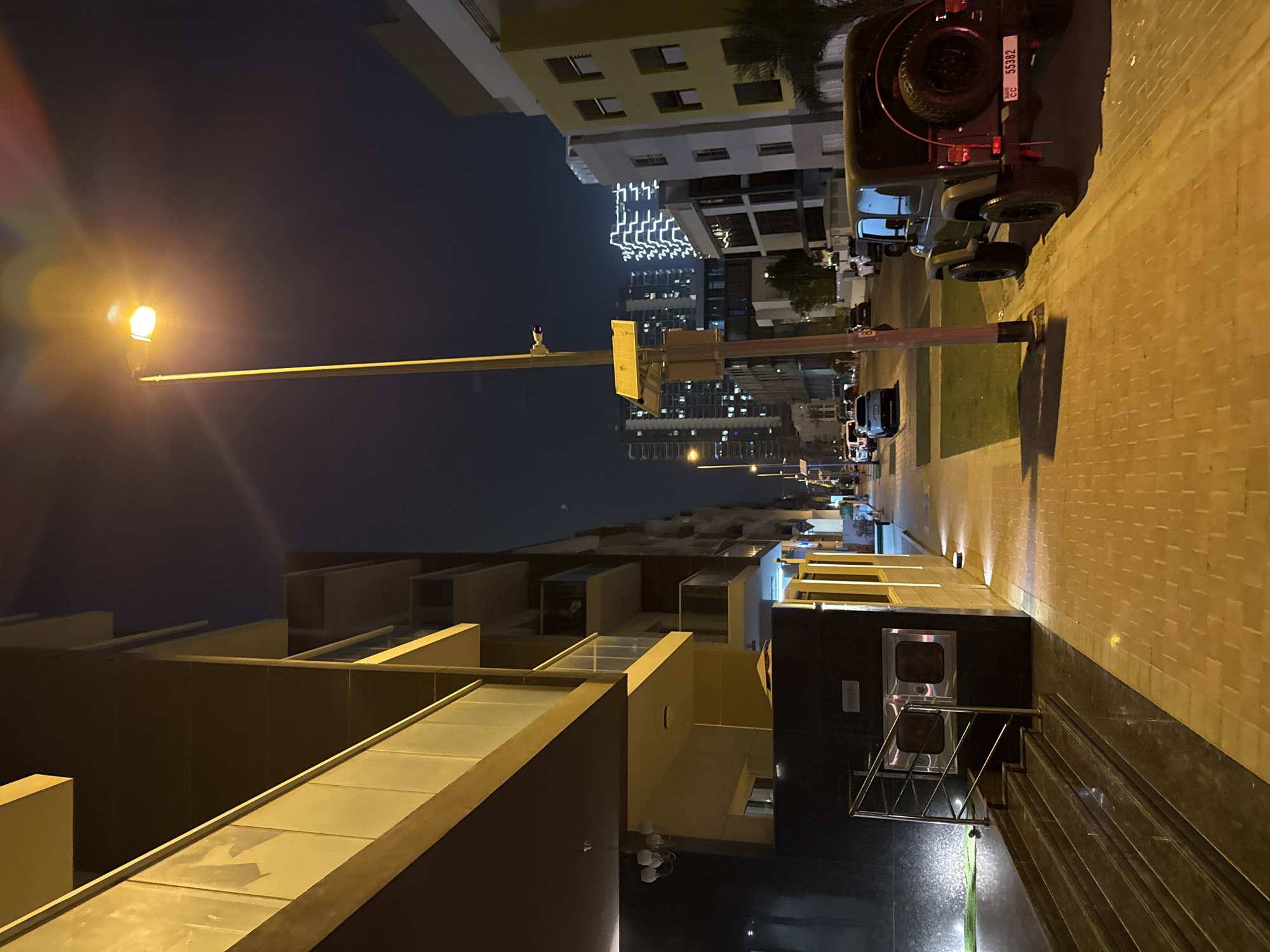 A street scene at night in Dubai, United Arab Emirates, features modern architecture and illuminated elements. The perspective is slightly tilted, with the street running from the bottom right to the top left of the frame.
On the right side of the image, a textured, light-colored brick sidewalk is illuminated by warm light. A dark-colored Jeep Wrangler with its spare tire visible is parked facing away from the camera, showing its license plate, which reads "CC 55382". Further down the street, other vehicles and the outlines of buildings can be seen.
On the left, a building with a dark facade and a sleek, modern design rises, featuring geometric balconies and large glass panels. A street lamp with a bright light source is prominently positioned in the mid-ground. In the distance, a cluster of tall buildings are illuminated with a distinctive pattern of white lights, suggesting a commercial or residential area. The sky is dark, indicating it is nighttime, and there are no clear indications of weather conditions. The overall mood is one of urban activity and modernity, typical of a city like Dubai at night.
