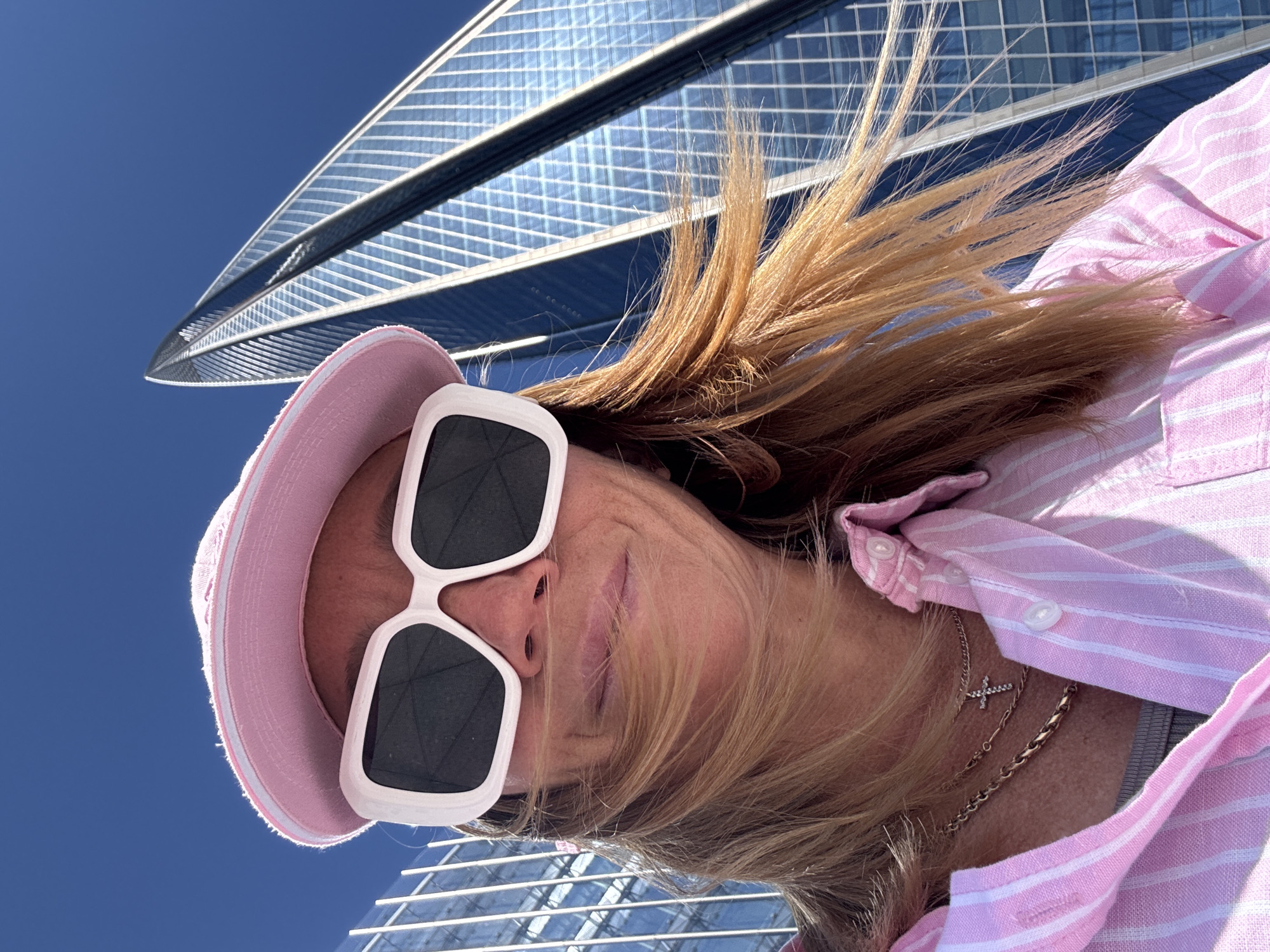 A low-angle selfie captures a person looking upwards, with a towering glass skyscraper in the background against a clear blue sky. The individual, appearing to be female, is positioned in the foreground, facing towards the sky. She wears a light pink bucket hat and distinctive white, square-framed sunglasses that cover her eyes. Her blonde or light brown hair is visible around her face, appearing swept back by the upward angle. She is dressed in a pink and white striped collared shirt, and two necklaces are visible around her neck, one featuring a small cross pendant and the other a simple chain.
The modern skyscraper in the background is a striking architectural element, characterized by its reflective glass facade and geometric grid patterns. The building appears to stretch high into the bright, unblemished blue sky, which suggests a sunny day with clear weather. The upward perspective emphasizes the impressive height of the structure. The scene depicts a casual outdoor moment in an urban setting, consistent with the context of Saint Petersburg, Russia. There is no visible text in the image.