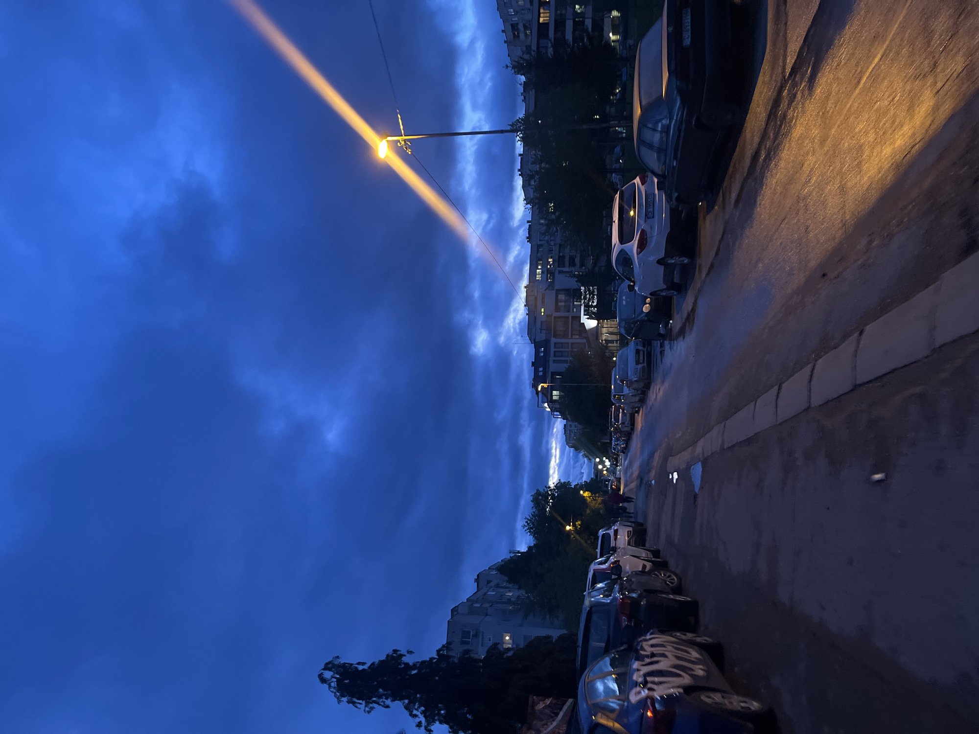 This image captures a street scene in Sofia, Bulgaria, at dusk or early night, looking down a residential street. The sky is a deep, dramatic blue, filled with prominent dark clouds and some lighter patches, particularly where artificial light interacts with them.
The central subject is a streetlamp, positioned on the left side of the street, which emits a powerful, yellowish beam of light upwards into the cloudy sky, creating a striking visual effect. The wet asphalt road surface reflects the artificial light, indicating recent rain or high humidity.
Numerous cars are parked along both sides of the street. In the foreground, on the left, there's a dark blue car with distinct white graffiti-like patterns on its side. Further down the street, a white car's license plate is partially visible, displaying "CB 9836 C*".
Multi-story residential buildings line both sides of the street, with several windows emitting warm light, suggesting occupancy. Trees are also visible, interspersed between the buildings and along the street, adding to the urban landscape. In the distance, on the right side of the street, a blurred figure, possibly a person, appears to be walking. The overall atmosphere is moody and atmospheric, characterized by the interplay of natural low light and artificial illumination.