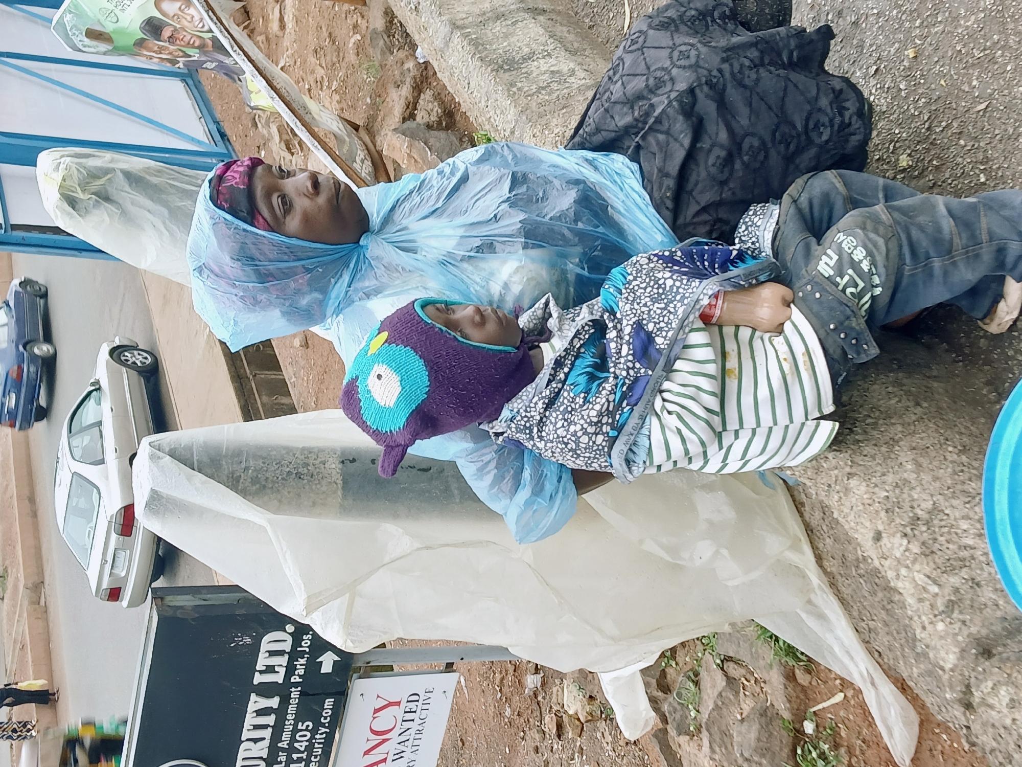 An outdoor scene shows a woman and a child lying on a concrete ledge, possibly resting or sleeping. The woman wears a floral head covering and is draped in a clear blue plastic sheet. The child, lying close to her, wears a purple knit hat and is covered with patterned fabrics.
The background reveals an urban environment. A sign is present bearing the words "JRITY LTD." and "Lar Amusement Park, Jos, 11405," placing the location in the city of Jos, Nigeria. The sign also features text indicating "VACANCY WANTED," suggesting a potential job opening.
Several vehicles are visible on the street, including cars.
The immediate surroundings include a white plastic sheet near the pair, along with exposed soil, some greenery, and a blue container near the edge of the ledge.