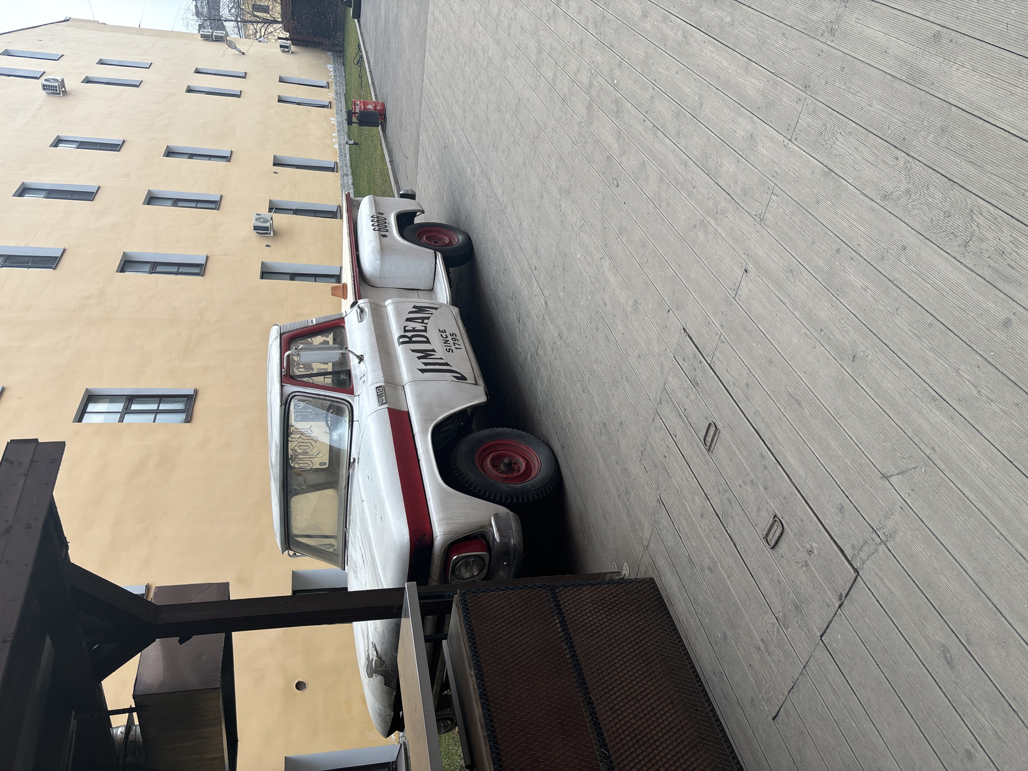 The image captures a vintage white and red Jim Beam truck parked on a wooden deck in what appears to be an urban setting. The truck, adorned with the Jim Beam logo "Since 1795," is parked parallel to a multi-story, light-yellow building with several windows and air conditioning units. The building suggests a commercial or residential structure.
A dark, metal structure, possibly part of an outdoor seating area or pergola, is visible in the foreground. The wooden deck is made of parallel planks, and a small patch of grass and a red fire extinguisher are visible further down the lane behind the truck. The scene suggests a blend of historical display and modern urban life.
Based on stylistic cues in the architecture and street furnishing, along with the Cyrillic script present in the car's interior, this photograph was likely taken in Saint Petersburg, Russia.
