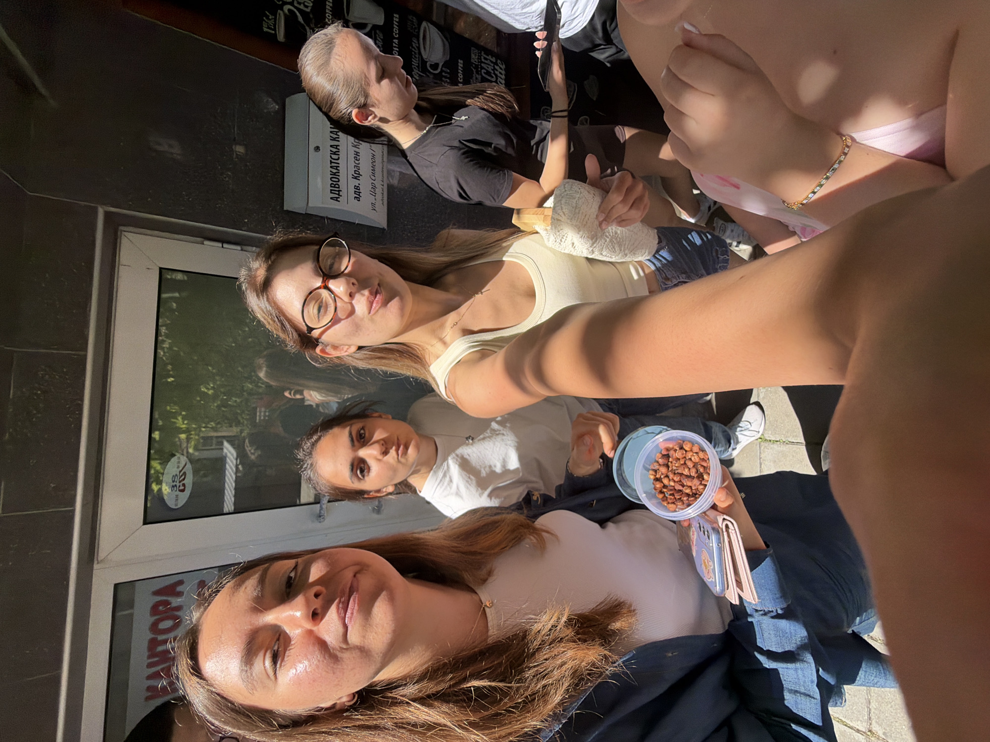 A group of four young women is captured in a selfie-style photograph outdoors on a sunny day in Burgas, Bulgaria. The primary focus is on the three women in the foreground and mid-ground.
On the far left, a woman with long brown hair, wearing a white tank top and a dark blue jacket, is looking towards the camera with a slight smile. She is holding a pink wallet in her left hand and a small, clear plastic container filled with nuts in her right hand. The container is partially obscured by her hand.
Next to her, a woman with dark hair and a white t-shirt is looking directly at the camera with a neutral expression.
In the center foreground, a woman with glasses and light brown hair is looking at the camera and smiling. She is wearing a light yellow tank top and holding a white cloth bag in her right arm.
Behind them, a fourth woman with her hair pulled back is wearing a black t-shirt and dark shorts. She is looking down and holding a smartphone in her left hand.
The background reveals the entrance to a building with a glass door. A sign with red lettering is visible on the door, possibly indicating a business name like "КАНТОРА" (Office). Another sign on the wall to the right has text including "АДВОКАТСКА КАНТОРА" (Law Office) and an address. The area appears to be a sidewalk with the pavement visible in the lower part of the image. The overall lighting suggests it is daytime with strong sunlight creating shadows.