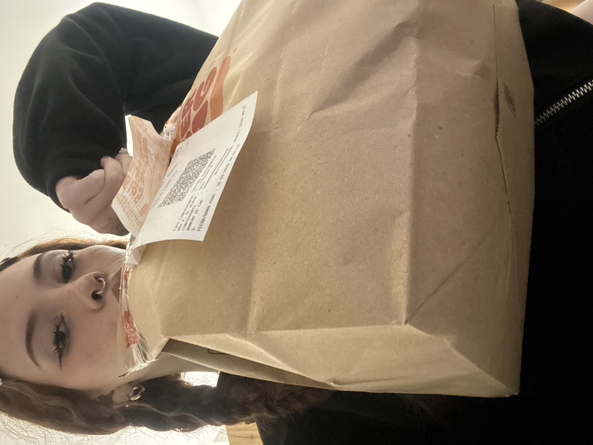 A person is holding a brown paper bag with a printed receipt attached. The person has fair skin, brown hair styled in a long braid, and visible facial piercings, including a nose ring and earrings. The paper bag has printed logos. The person wears a black sweatshirt and is indoors in a dimly lit area. The location is Buenos Aires, Argentina.