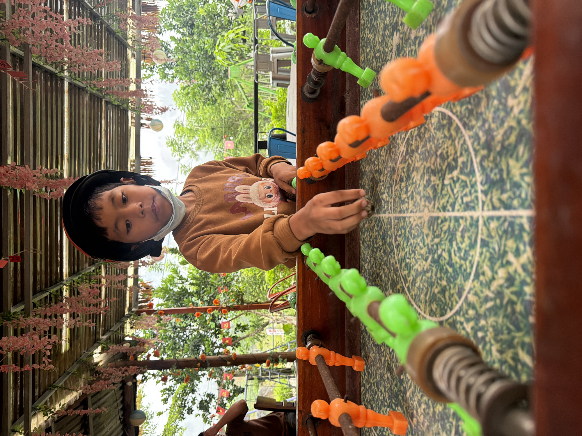 A young child wearing a black beret and a light blue disposable face mask is playing foosball at what appears to be an outdoor cafe in Đà Nẵng, Vietnam. The foosball table has orange and green players. The playing field has a green and white pattern with a white circle in the center. The cafe has a wooden ceiling with pink hanging decorations. In the background, there are trees, some decorated with small Vietnamese flags, and some metal chairs. A person's legs are visible in the lower left corner.