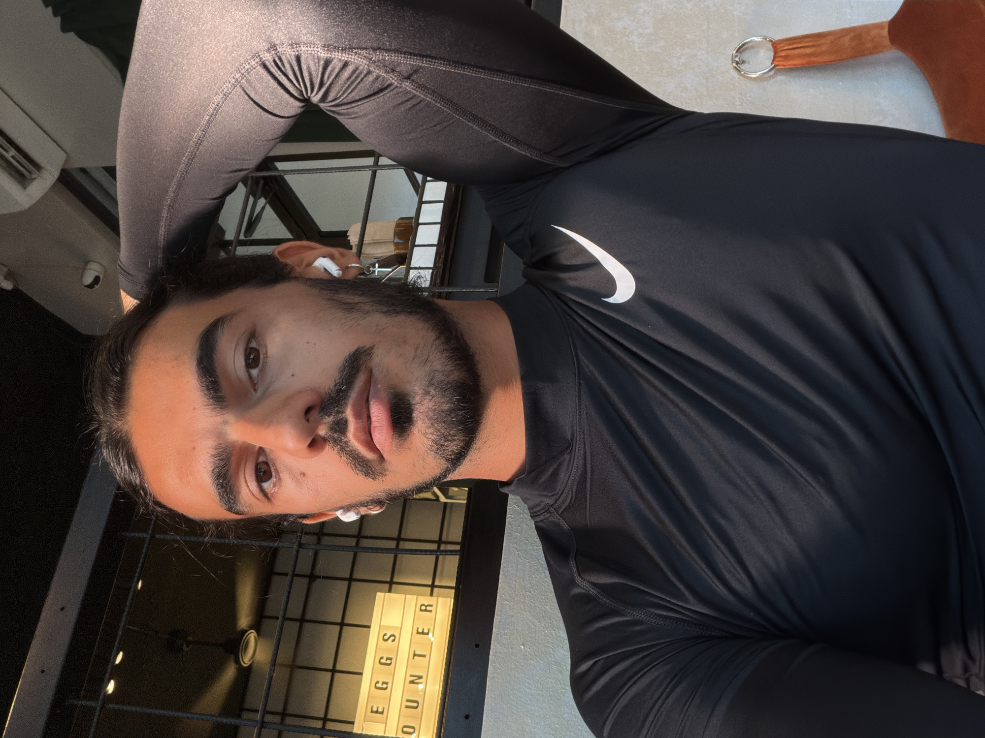 A man with dark hair and a beard is wearing a black Nike athletic shirt and leggings. He is facing the camera with an earbud in each ear. Behind him, a sign reads "EGGS COUNTER." The location is North Kuta, Indonesia.