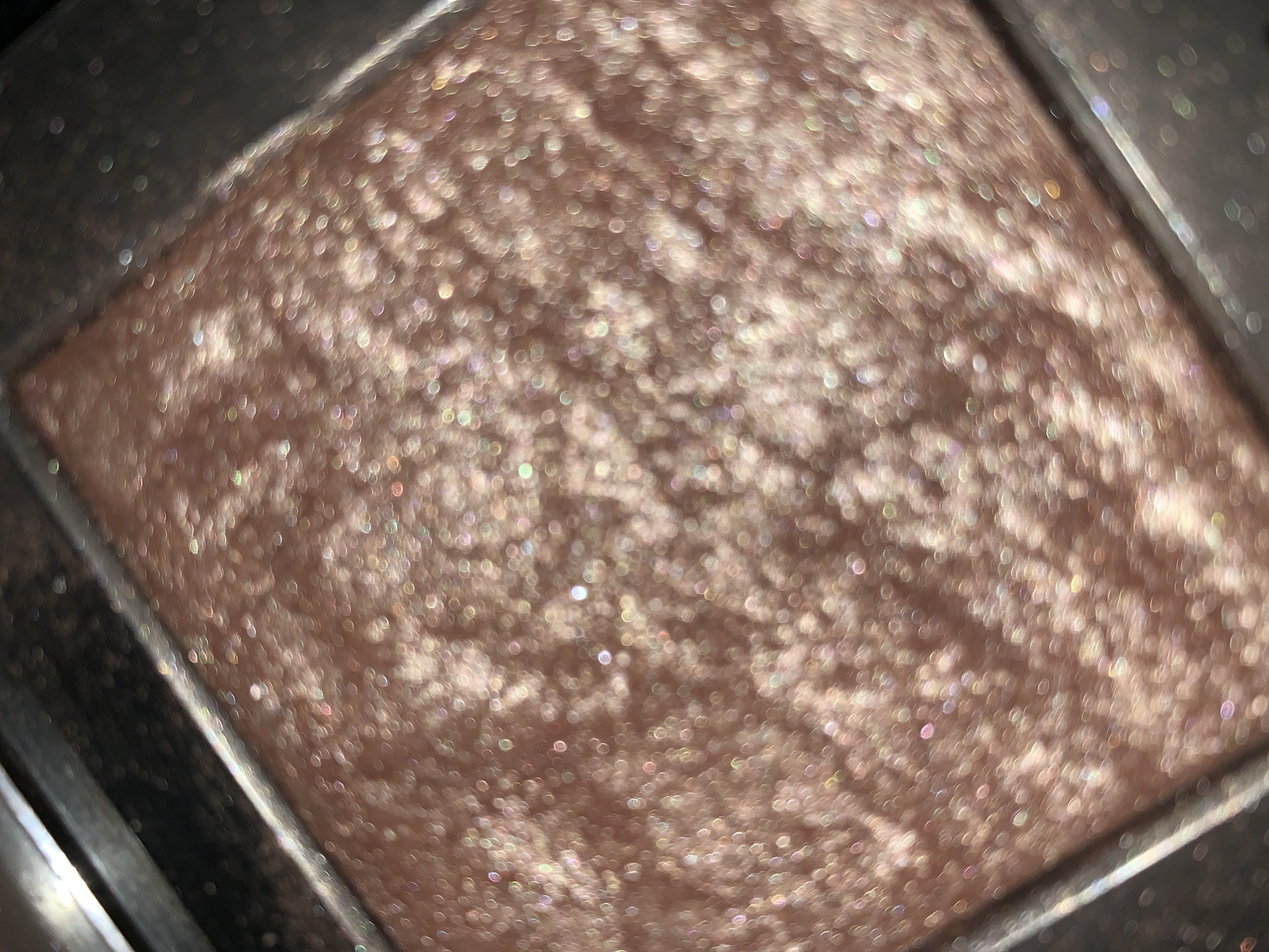 A close-up, out-of-focus image shows a square of makeup in a container. The makeup is a light pinkish-tan color with a glittery or shimmery texture. The container appears to be a dark gray or black color. The location and specific city are unknown.