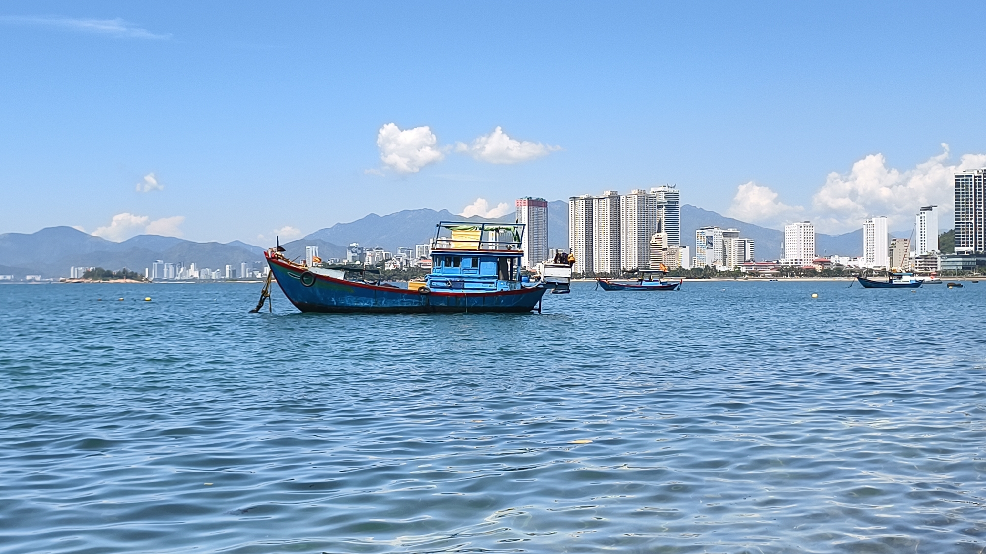A bright, sunny day at the coast of Nha Trang city, Vietnam, is captured in this image. The foreground is dominated by rippling blue water, with a light reflection creating a luminous effect. In the mid-ground, a blue and red fishing boat is anchored, its rope taut and disappearing into the water. This boat is well-equipped with a cabin structure and various poles. Further out, a second, smaller dark-colored fishing boat is visible.
The background features the Nha Trang skyline, a collection of modern high-rise buildings of varying heights and designs, predominantly white. Behind the city, a range of majestic, hazy blue mountains forms a dramatic backdrop. The sky is a clear, bright blue, punctuated by a few fluffy white clouds. The overall impression is one of a peaceful coastal urban landscape under a clear sky.
