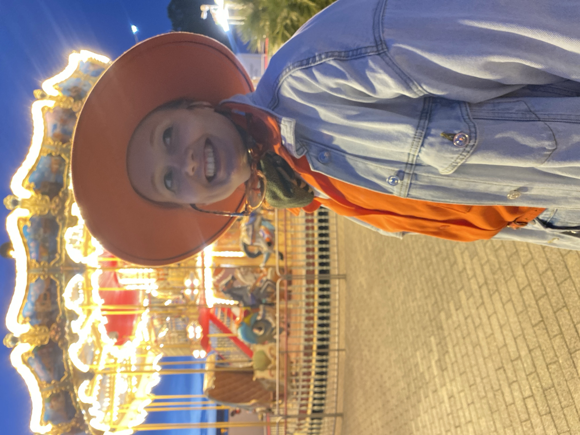 A person wearing an orange hat, orange shirt, and denim jacket is standing in front of a carousel. The carousel is brightly lit with golden lights and is detailed with ornate decorations. The ground is paved with beige bricks. The image was taken at night in Sochi, Russia.