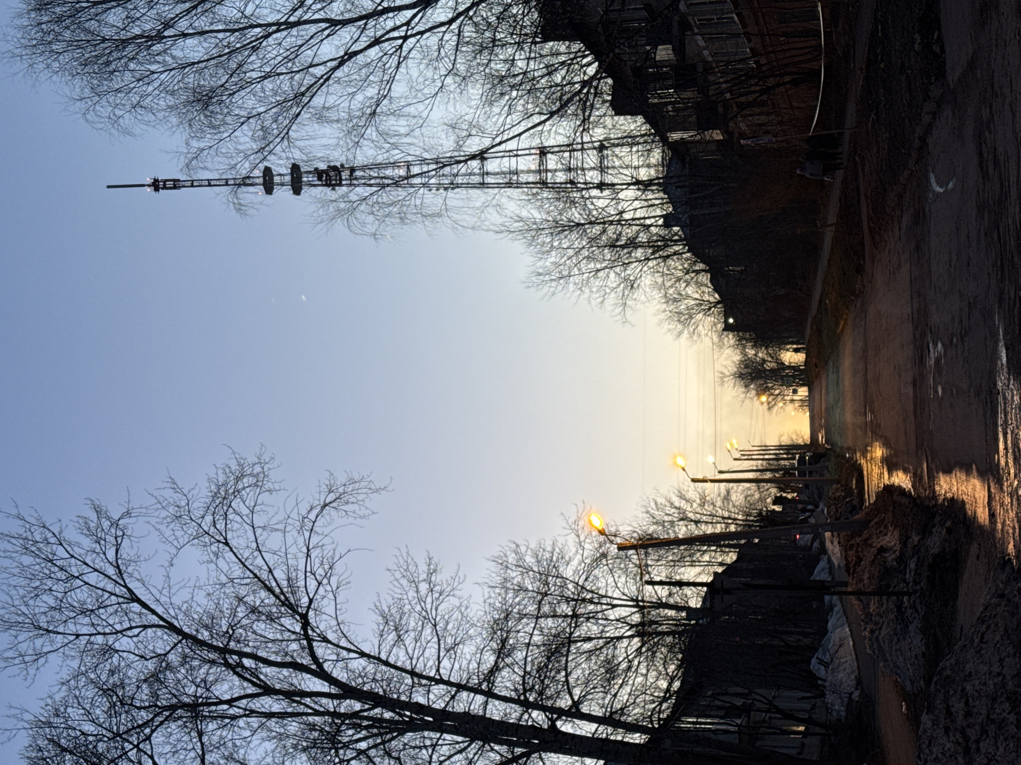A low-angle shot of a street in Berezniki, Russia. The street is flanked by bare trees and buildings silhouetted against a twilight sky. Streetlights illuminate the road. A tall radio tower dominates the skyline on the left. A layer of snow or slush is present on the ground.