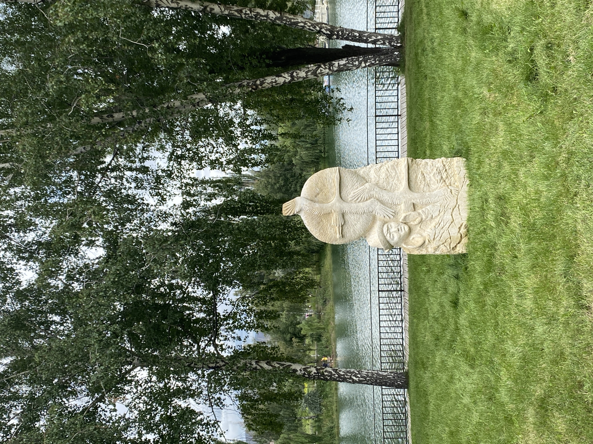 The image depicts a stone sculpture situated on a lush green lawn in Cholpon-Ata, Kyrgyzstan. The sculpture is carved in a vertical orientation and features a human face with distinctive headwear on its lower portion, above which is a relief of a bird with outstretched wings. In the background, a calm body of water, likely Lake Issyk-Kul, is visible, bordered by a black metal fence and a wooden walkway. Several trees with white bark and green leaves, possibly birch trees, are prominent on either side of the sculpture, casting shadows on the grass. Further in the distance, a verdant treeline stretches across the horizon, with mountains faintly visible beyond. The sky is overcast with clouds. No individuals are actively engaged in any interactions within the frame, though a few figures can be seen near the water's edge in the distance.