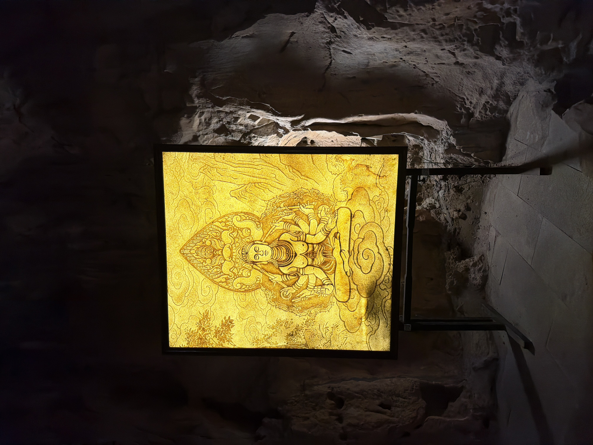 The image depicts a square, illuminated panel affixed to a rough, rock-like wall, possibly inside a cave or grotto. The panel features a golden, intricate illustration of a deity, possibly a Buddhist figure, seated on a cloud or lotus. The wall's texture suggests a natural, geological formation, with crevices and uneven surfaces. The overall lighting is dim, except for the panel's bright illumination, creating a contrast. The panel is mounted by what appears to be a dark metal bracket. The scene is potentially located in the Marble Mountains of Đà Nẵng, Vietnam, given the grotto-like environment and religious iconography, but that cannot be stated with absolute certainty based on the image alone.