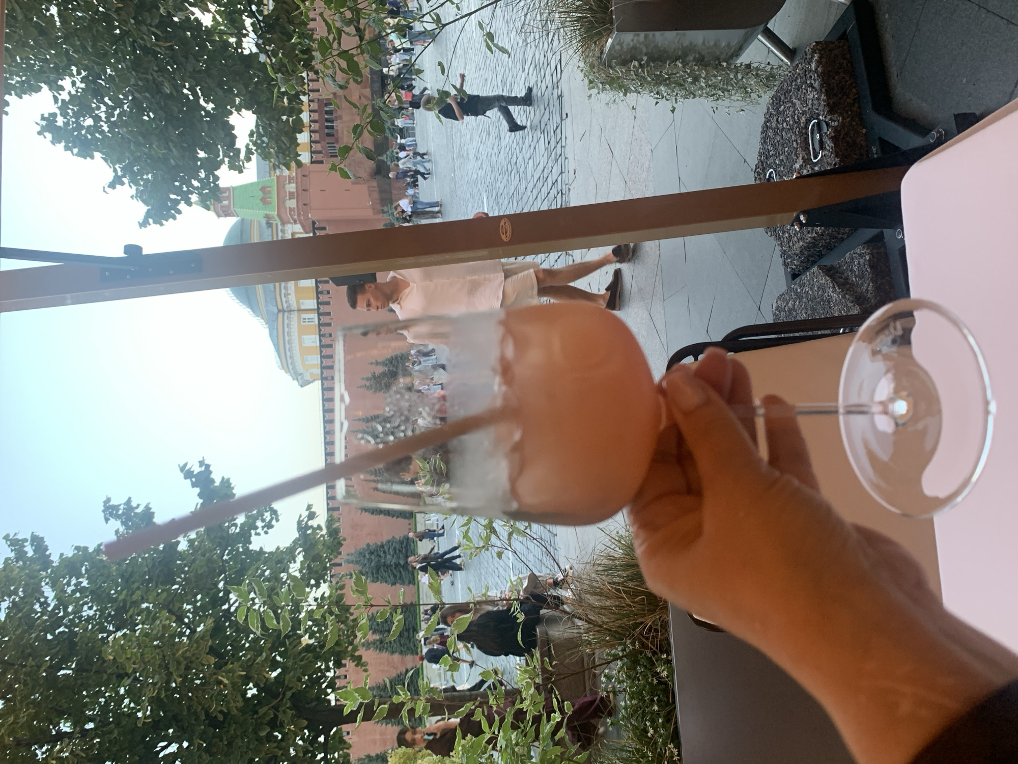 In the foreground, a hand holds a wine glass filled with a pink beverage and ice, with a straw. Behind the glass, a man in a white short-sleeved shirt and light shorts is walking. In the background, the red brick wall of the Kremlin and several trees are visible, with people walking on the cobblestone ground. The setting appears to be an outdoor seating area in Moscow, Russia, likely near Red Square, given the prominent Kremlin architecture. The sky is clear and bright, suggesting daytime.