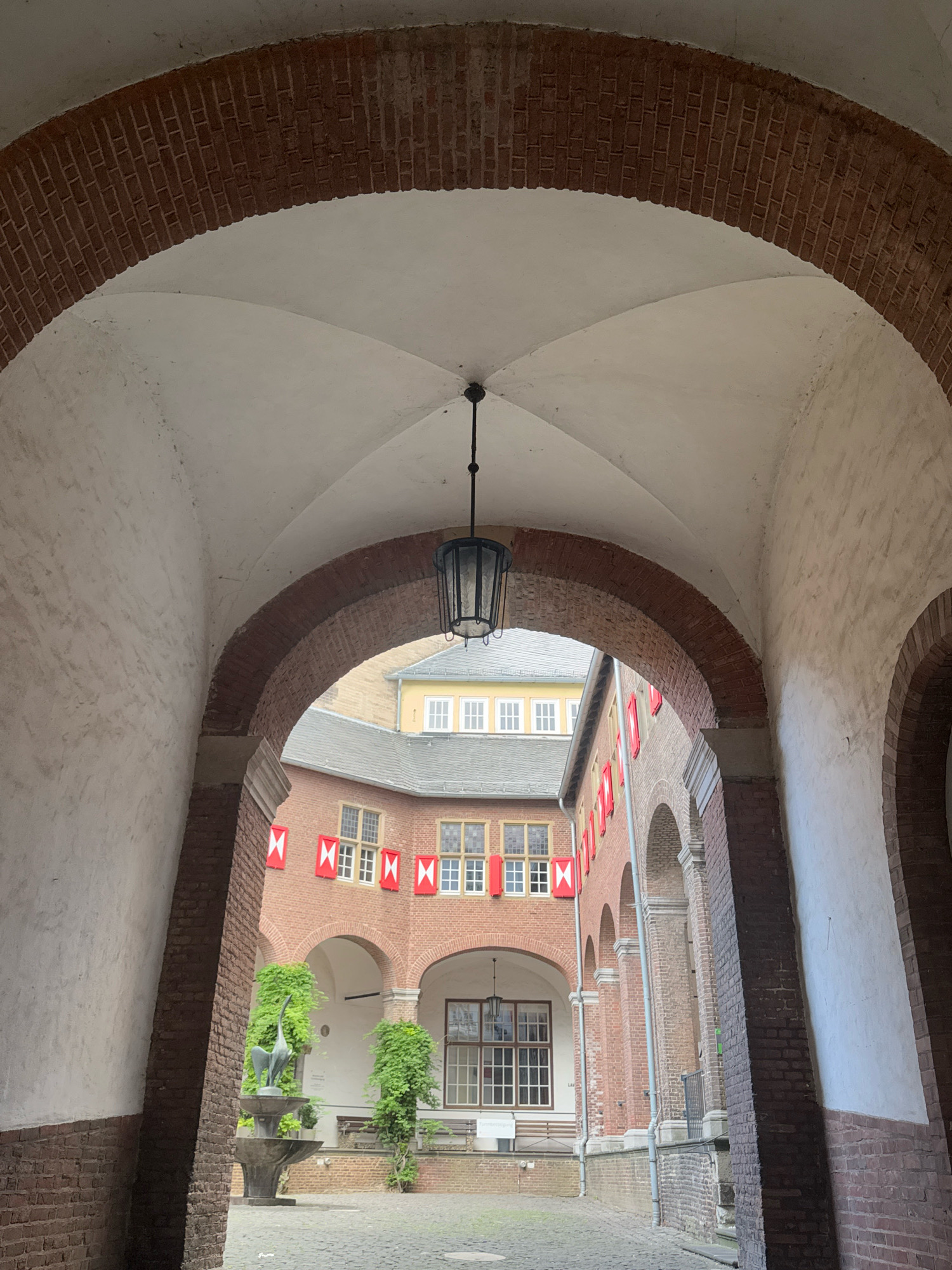 This image depicts the inner courtyard of the Cleves Castle (Schloss Kleve) in Cleves, Germany, as viewed from a brick archway. The courtyard features red brick buildings with arched windows, some adorned with red and white shutters. A fountain with a bird sculpture stands prominently in the center of the courtyard, surrounded by greenery. A hanging lantern is suspended from the vaulted ceiling of the archway. The ground is paved with cobblestones.