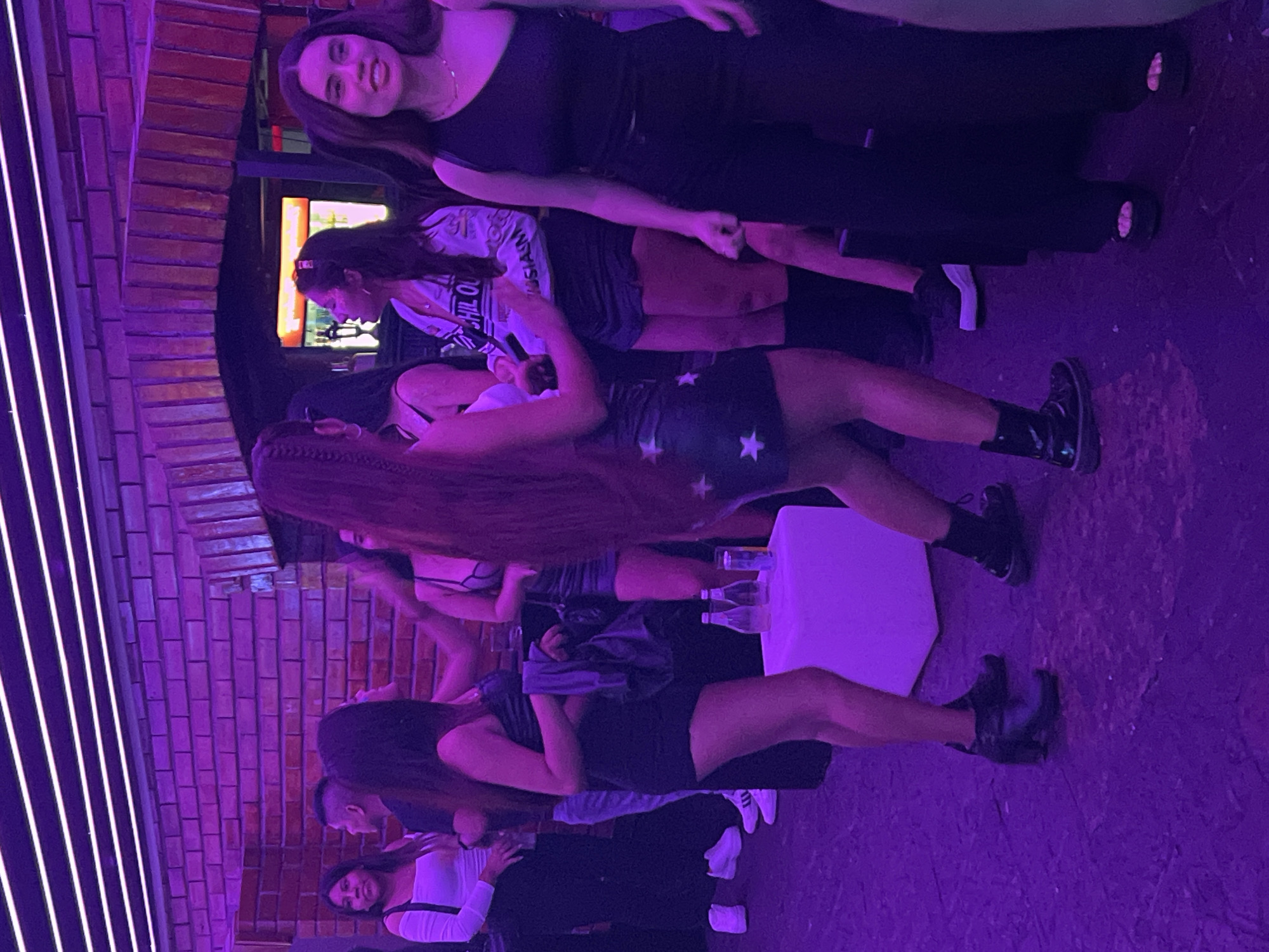 The image shows a group of women in a dimly lit indoor space, likely a nightclub or bar. The lighting has a strong purple hue. A brick wall with decorative neon lights is visible. There is a small white table with bottles and glasses on it. Most of the women are wearing short dresses or skirts and dark footwear. One woman near the top of the frame is wearing a long black dress. The location of the photo in the city of Arica, Chile is not supported by the image alone.