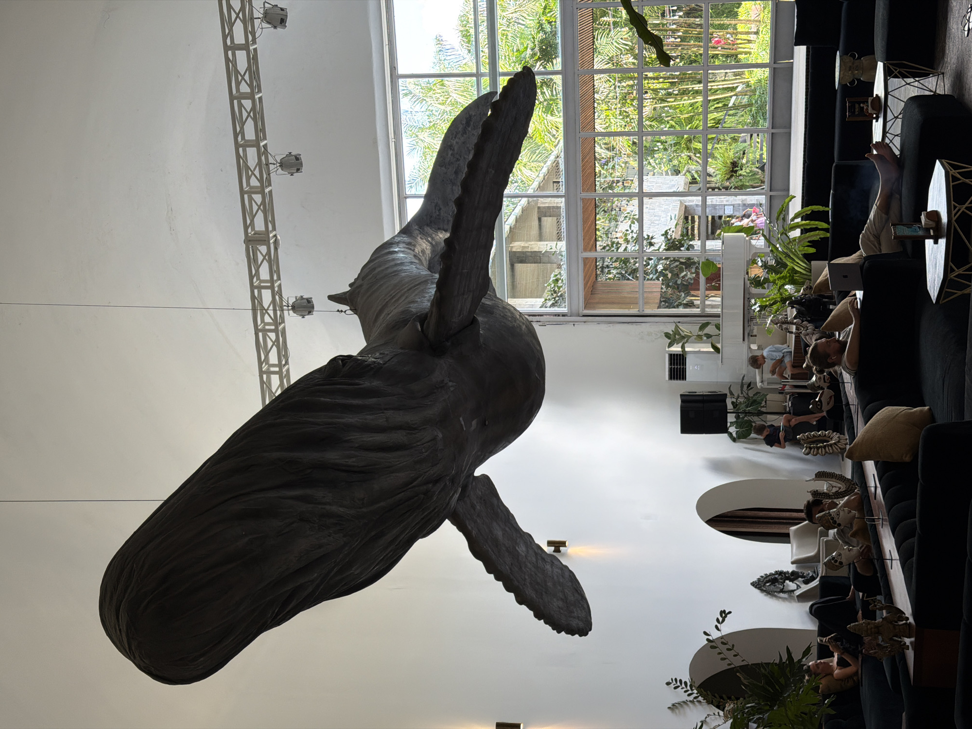 The image shows the interior of a large, modern space, likely a lounge or lobby, with a prominent, dark sculpture of a whale suspended from the ceiling. The whale sculpture is detailed and appears to be made of a textured material. The space features large windows that look out onto a lush, green outdoor area with palm trees. Inside, there are several people seated on dark sofas and chairs, some engaged with their phones or laptops. A white grand piano is visible in the background, with two children nearby. The architecture includes white walls, arched doorways, and a system of industrial-style lighting fixtures. The overall atmosphere is contemporary and relaxed, with a blend of indoor comfort and natural elements visible through the windows. The scene is located in the Ubud District, Indonesia.