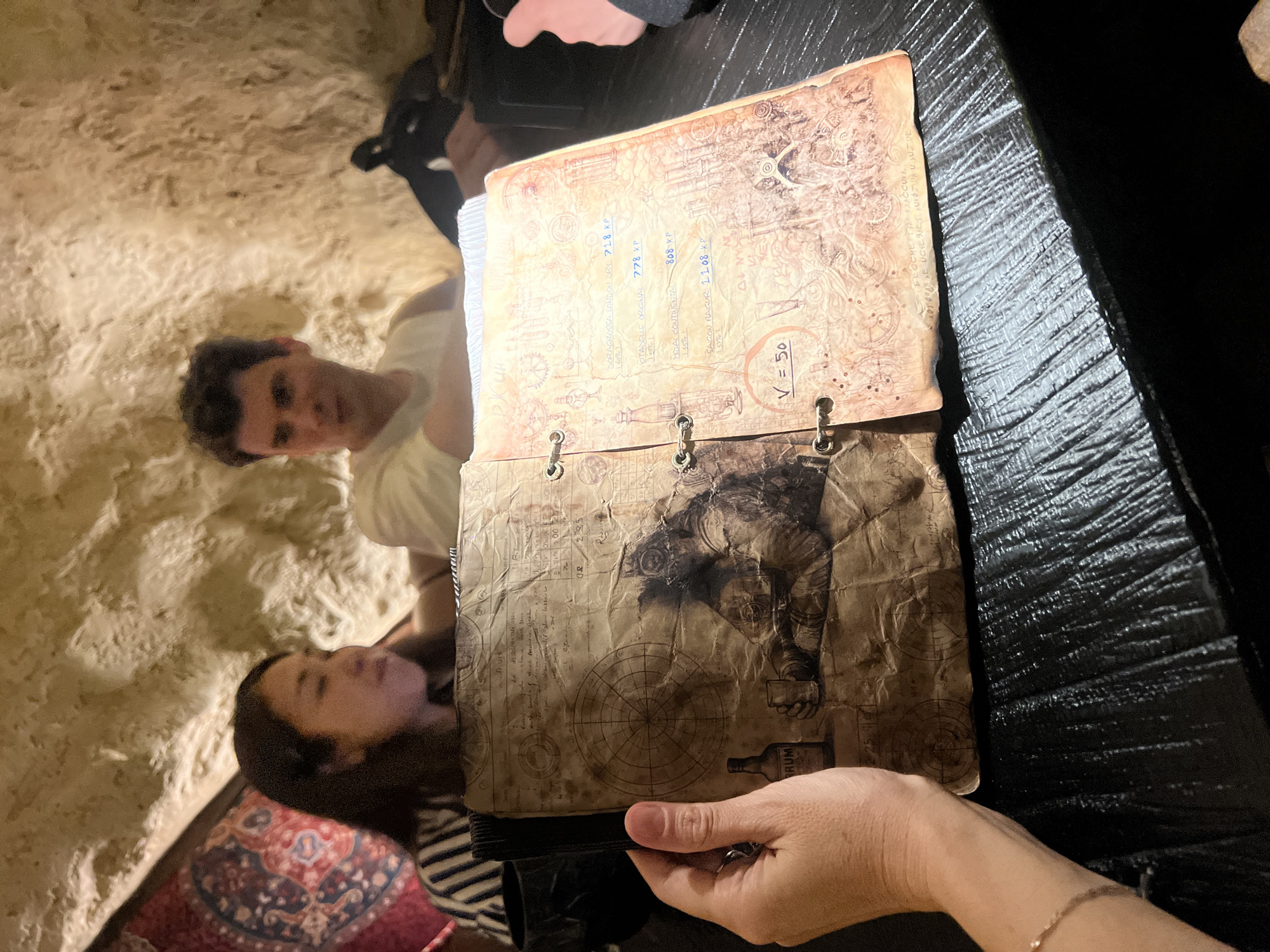 The image depicts an interior scene at the Karakum Bar in Moscow, Russia, characterized by its unique cave-like ambiance and adventure-themed decor. In the foreground, a person's light-skinned hand, adorned with a delicate bracelet and a silver-colored ring on the ring finger, holds open a dark-bound menu designed to resemble an ancient, weathered parchment logbook or map.
The menu's left page features a large central circular diagram akin to a compass rose, accompanied by a detailed illustration of a figure (possibly an old-time diver or adventurer) in heavy gear, sitting with a glass and a bottle explicitly labeled "RUM." Various handwritten notes and smaller diagrams are also present on this page. The right page of the menu lists several beverage options with their prices in "KP," including "DORAGROSSA LONDON DRY 718 KP (50%)," "CITADELLE ORIGINAL 778 KP (50%)," "HIDAI CONTINENTAL 868 KP (50%)," and "SAGON NAVIGUR 1108 KP (50%)." Below these items, "V = 50" is written, and the page is decorated with intricate borders and faint background sketches.
The menu rests on a dark, textured table surface that appears slightly reflective. In the mid-ground, a woman with long dark hair, wearing a striped top, is visible on the left, looking downwards, her face partially obscured. To her right, a man with short dark hair, wearing a light-colored t-shirt, looks towards the left with a neutral expression. A partial view of another person's arm in a dark sleeve is visible on the far right.
The background features rough, irregular, light-colored walls, mimicking natural rock or a cave interior, consistent with the thematic design of the Karakum Bar. A patterned tapestry or fabric in red, blue, and white is visible on the upper left portion of the background wall. The lighting is dim and ambient, contributing to a cozy and mysterious atmosphere. The currency "KP" (Karakum Points), the "RUM" illustration, and the cave-like setting firmly establish the location as the Karakum Bar in Moscow, Russia.