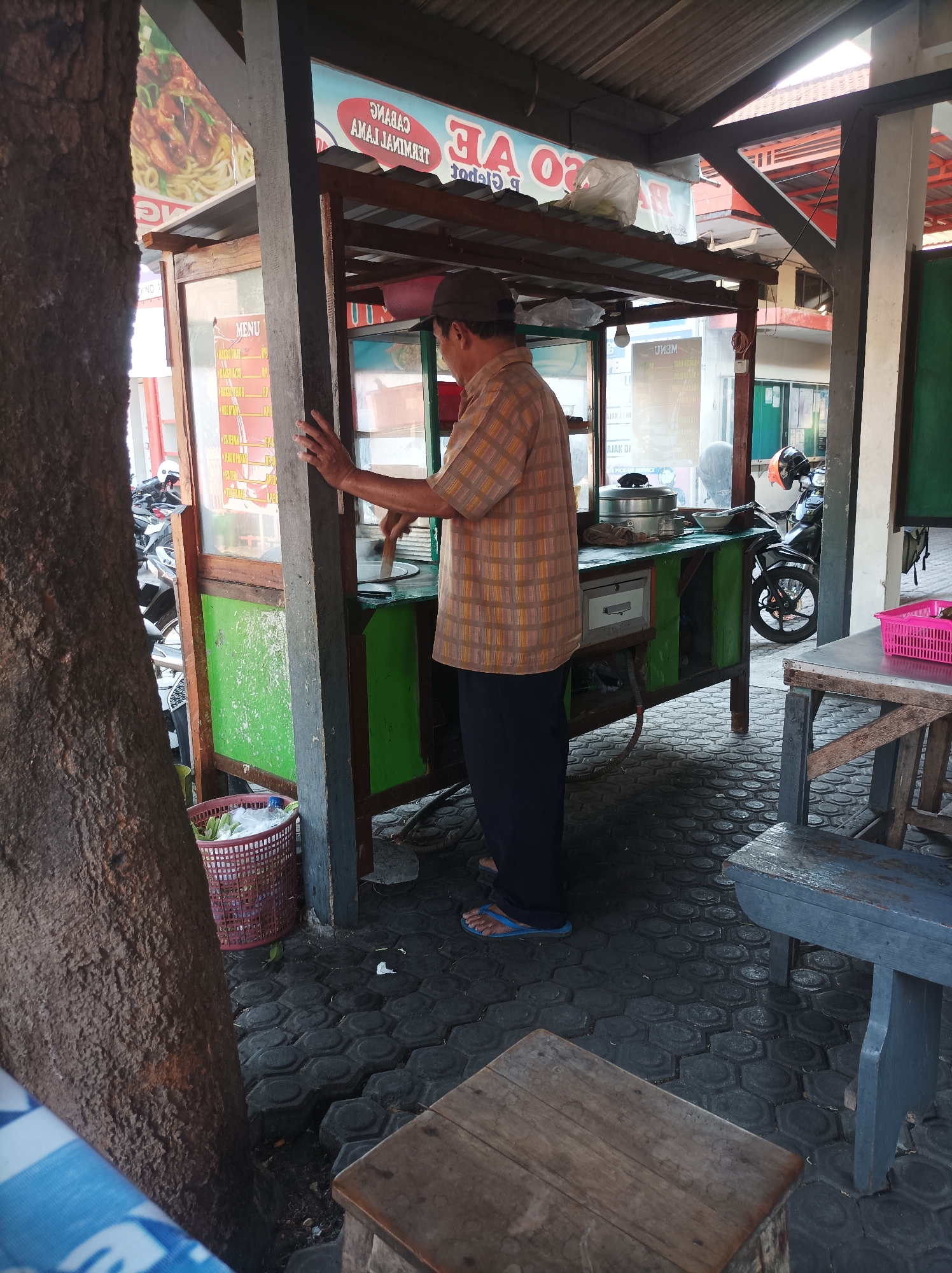 A man is seen from the back, wearing a plaid shirt and dark pants, standing at a food stall in Nganjuk, Indonesia. He appears to be cooking or preparing food, with his hands reaching towards a circular pan or griddle. The food stall is made of wood and painted green, with a glass display case showing a menu. A red woven basket filled with plastic bags is placed on the ground near the stall. In the background, motorcycles are parked, and the scene is partially shaded by an overhang. The ground is paved with hexagonal dark gray tiles.
