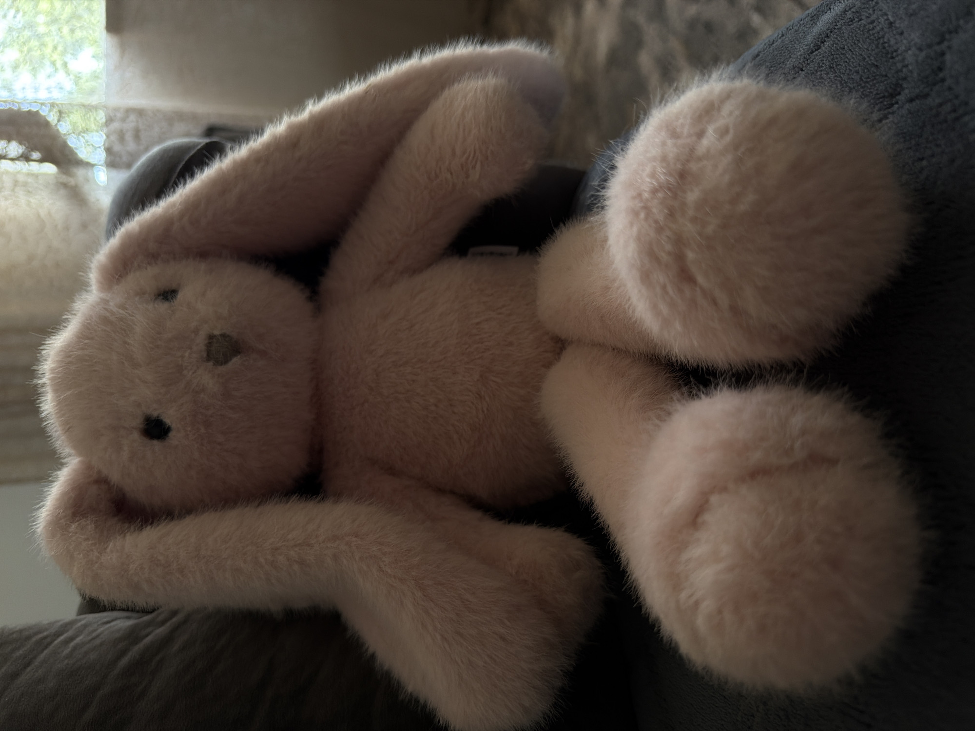 A plush pink bunny toy is seated on a dark gray sofa. The bunny has long, floppy ears, small black eyes, and a pink stitched nose and mouth. Its legs are extended forward, and its arms are resting at its sides. The background is blurred, showing a window with sheer curtains and greenery outside, suggesting an indoor domestic setting. The location is an unspecified area within Lernamerdz, Armenia.