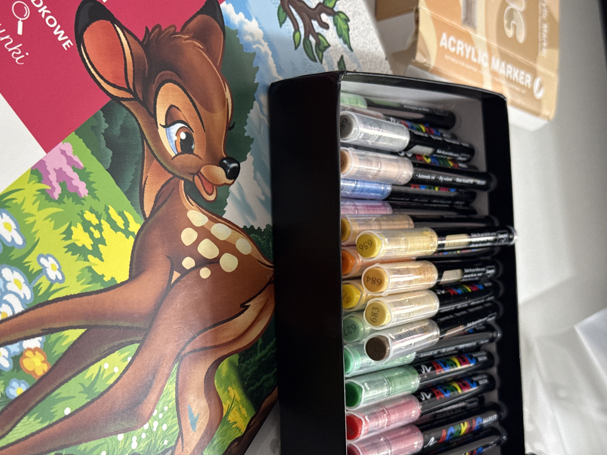 The image shows a collection of art supplies, primarily marker pens, neatly organized in a black case. The case is positioned to the right of a colorful illustration featuring Bambi, the Disney character, set against a backdrop of lush green foliage and white and yellow flowers.
The Bambi illustration appears to be part of a book or poster, with the words "Dkowki" and "nki" visible in white text on a pink background in the upper left corner. Another book or package with the text "ACRYLIC MARKER" in large, bold letters is partially visible in the upper right corner.
The marker pens in the case are of various colors, with clear plastic barrels allowing the ink colors to be seen. Some of the markers have numerical codes printed on their caps, such as "959," "684," "683," and "627." The markers appear to be brand new or well-maintained, suggesting they are ready for use.
The overall scene conveys a sense of creativity and artistic endeavor. The bright colors of the illustration and the organized markers imply that someone is either preparing to create art or has recently finished a project. The setting appears to be indoors, possibly a desk or workspace, given the presence of art supplies and printed materials. The lighting is bright, suggesting daytime. No specific time of day or weather conditions can be determined. The location context of Łódź, Poland, is provided but not visually evident in the image itself.