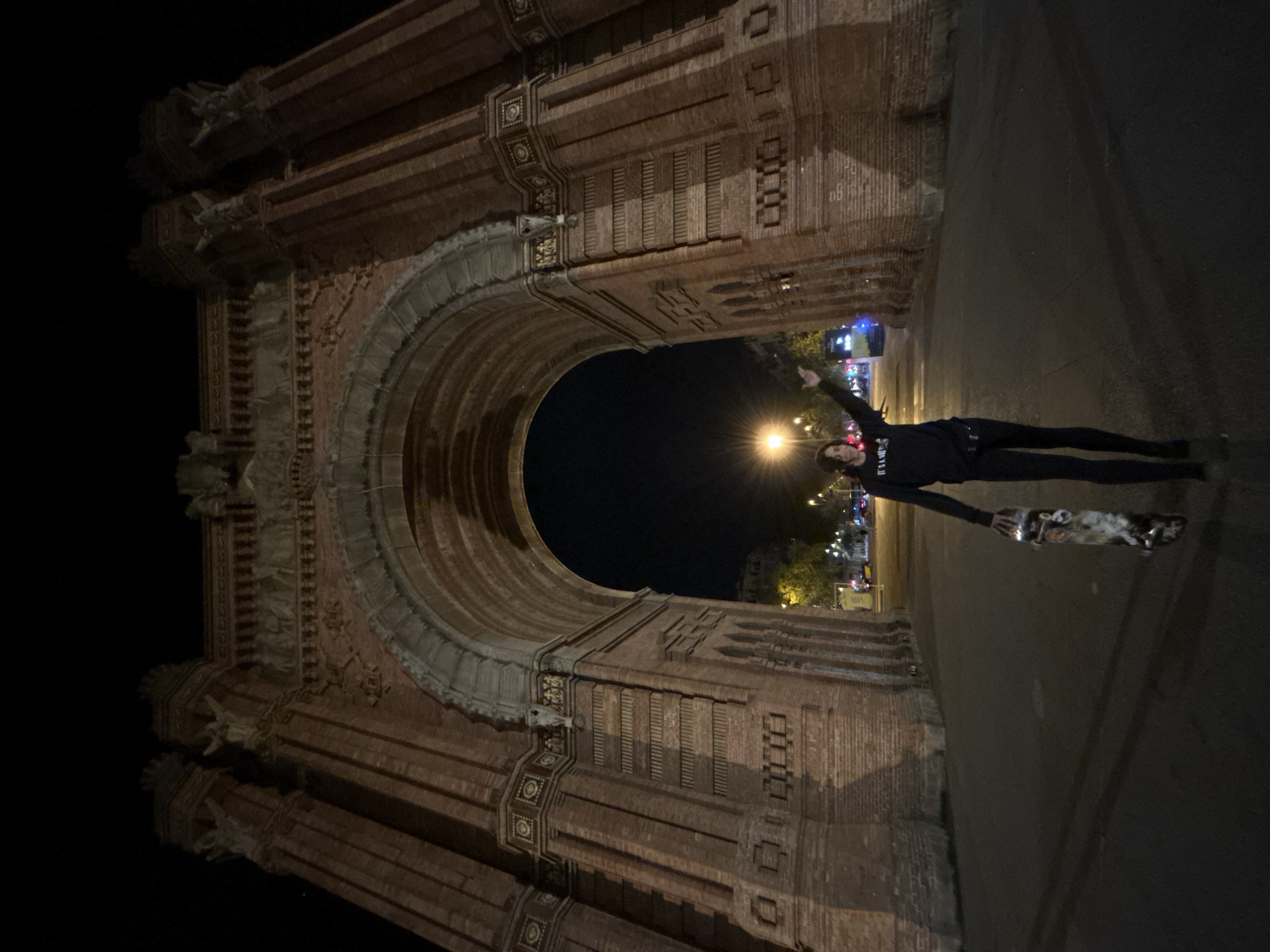 A horizontal photograph taken at night shows a person holding a skateboard in front of the Arc de Triomf in Barcelona, Spain. The arch is illuminated and its brick facade is visible. The person is wearing dark clothing and holding one arm up. Streetlights are visible in the background.