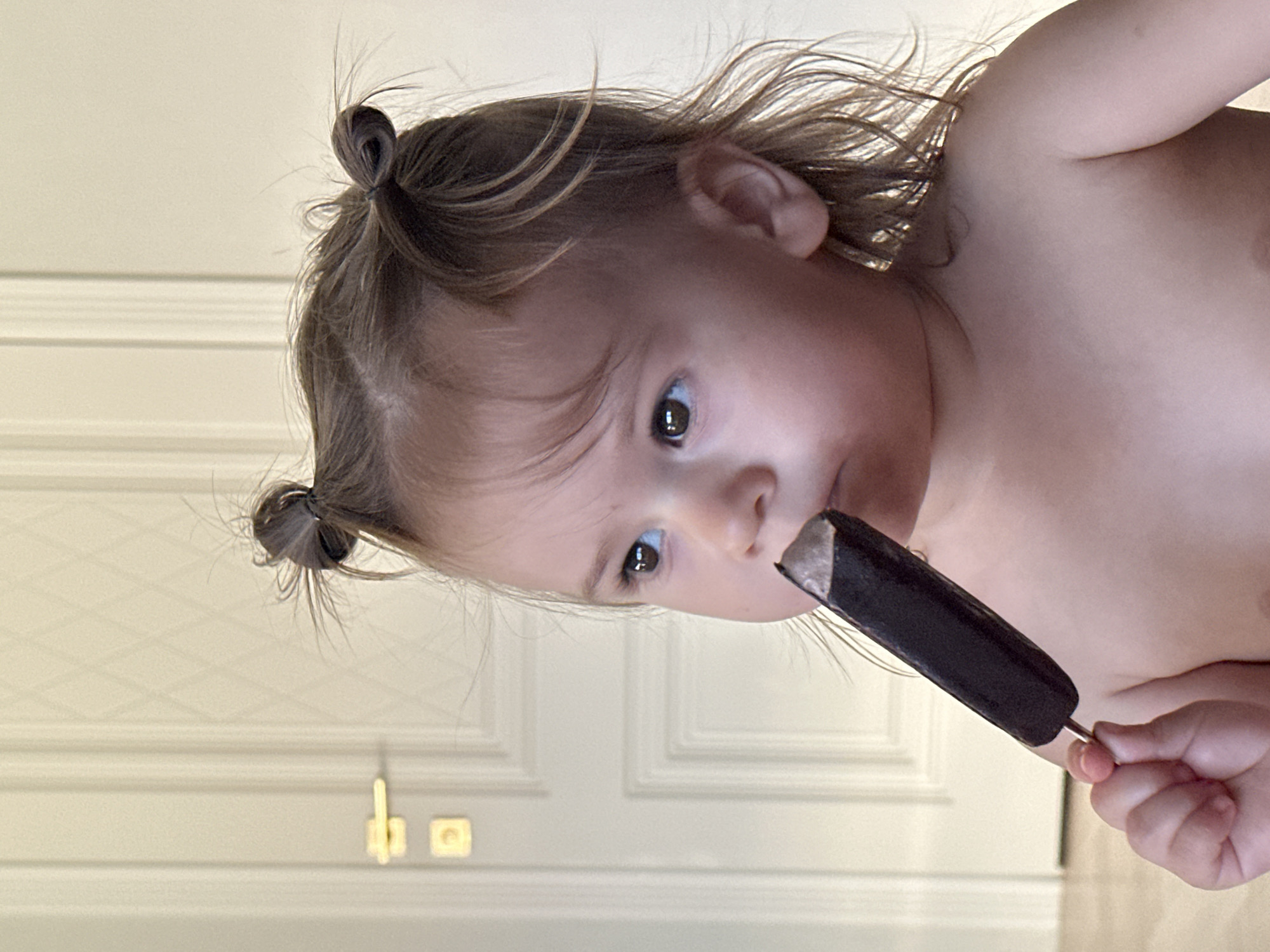 A young child with light skin, dark eyes, and brown hair styled in two small buns, is holding a dark-colored popsicle. The child appears to be indoors, with a white door featuring recessed panels and gold-colored hardware visible in the background. The photograph is taken from an unusual angle, with the child positioned in the upper right and the door occupying much of the lower left of the frame. There is no evidence of the location being in Kharnavy, Russia.