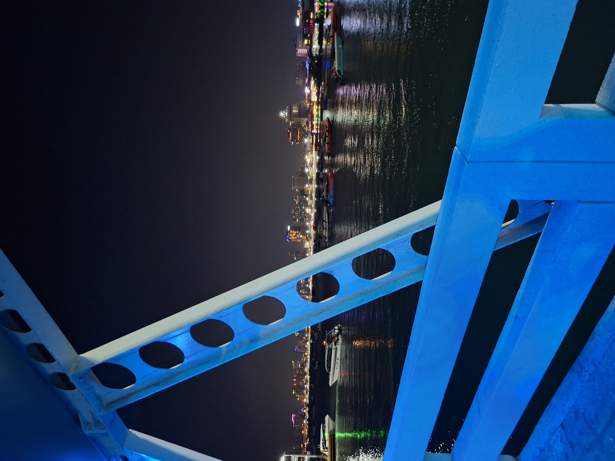 The photograph depicts an out-of-focus nighttime view of Pattaya City, Thailand, seen from a high vantage point, presumably a bridge structure. The city skyline is visible along the waterfront, illuminated by bright artificial lights that reflect off the water's surface. Several boats, also lit, are visible on the water.
The foreground of the image features a blue-lit structural element, likely part of the bridge, which is out of focus and abstracting the view of the city and water. This structure is made of white metal beams, some with circular cutouts, giving the photograph an architectural and urban feel. The rest of the sky is dark.