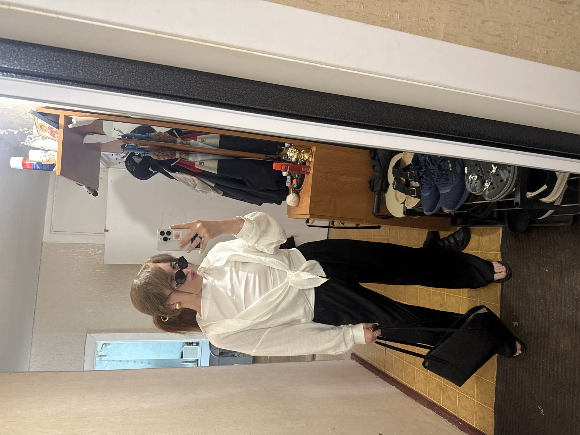 A medium shot shows a person facing left and holding a smartphone in their right hand, likely taking a selfie in a mirror. The person is wearing sunglasses, a white long-sleeved top knotted at the waist, and black trousers, and holding a black purse. A ponytail is visible. The reflection reveals a hallway cluttered with items on shelves, including shoes, umbrellas, and other stored goods. The floor is tiled in a light brown pattern. The location appears to be a domestic interior, possibly in Chaikovsky, Russia, as specified.