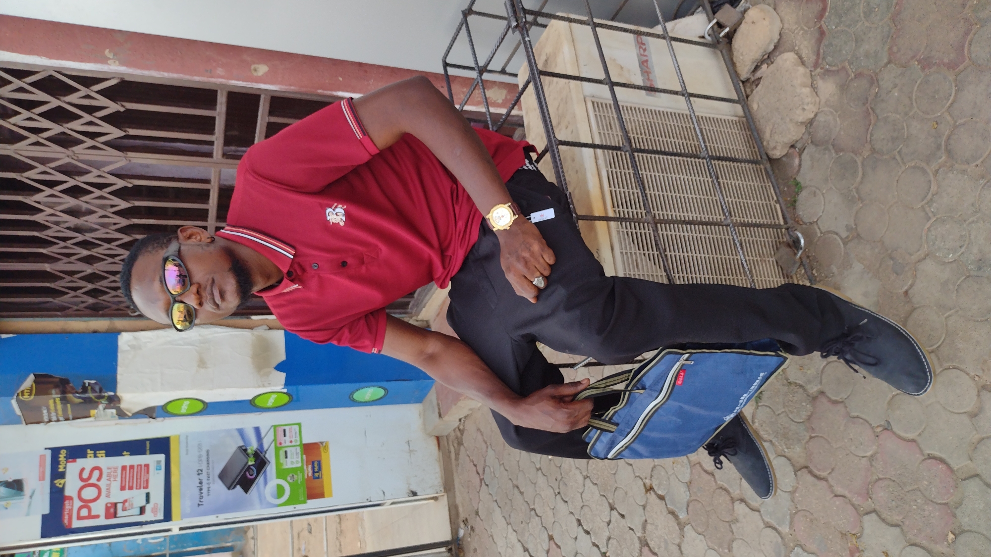 A man stands on a paved area. He wears a red polo shirt with a small white emblem on the chest, black pants, and black shoes. He also has sunglasses on and a gold watch on his left wrist. He is holding a blue bag in his right hand. Behind him, there is a building with blue and white signage, and a metal security gate. There is a Sharp brand object attached to the building wall as well. The location is in Jos, Nigeria.
