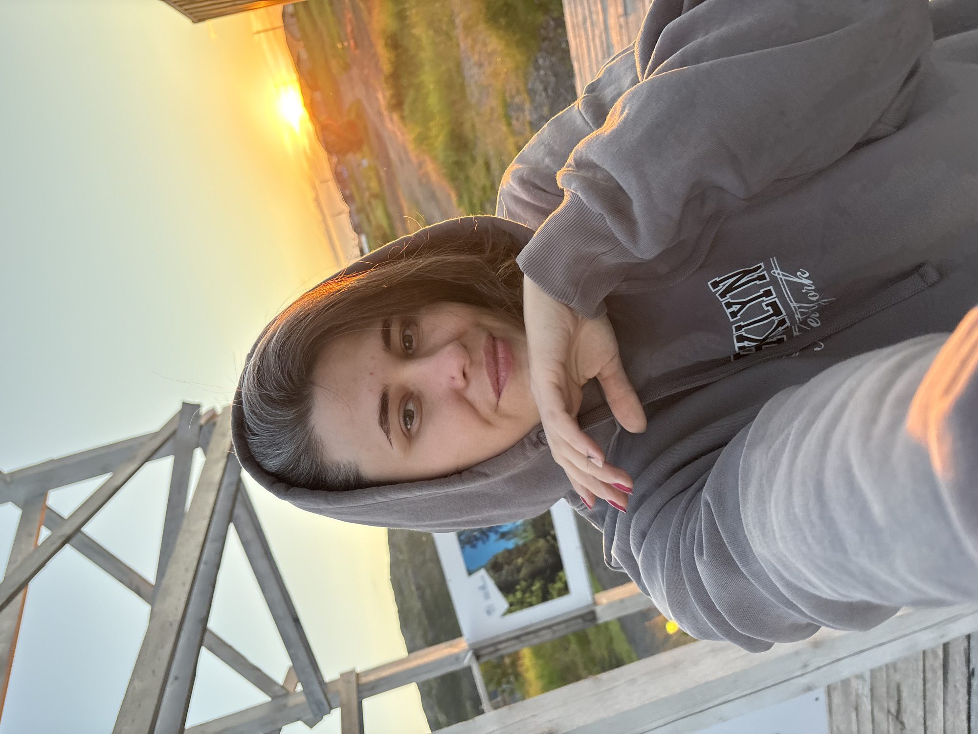 A woman with short, dark hair and streaks of gray is taking a selfie outdoors. She is wearing a gray hooded sweatshirt with the word "Brooklyn" and "New York" printed on it in white lettering. She is leaning her chin on her hand, with her nails painted bright pink. The background shows a sunset over a body of water and a small town. To the left of the woman is a wooden structure, possibly a gazebo or shelter, and a framed picture on a white board. The scene appears to be in Teriberka, Russia, a coastal village.