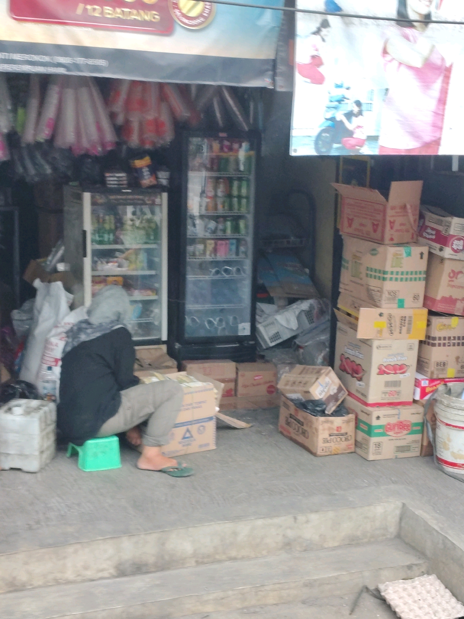 The image depicts an outdoor scene in front of a small shop in Tangerang, Indonesia. The shop appears to be a general store or minimarket, as indicated by the display refrigerators filled with drinks and snacks, and the stacks of boxed goods outside. In the foreground, a person wearing a grey hijab and a dark sweater is crouched on a small green stool, facing away from the camera and towards a stack of boxes. They are wearing light-colored pants and flip-flops. To the left of the person, there is a large white plastic container.
The shop's facade features a large banner at the top with text, likely advertising products or the shop's name. Below this, several items are hung, possibly plastic bags or other merchandise. The refrigerators inside the shop are visible, showing various products on their shelves.
Outside the shop, the ground is paved, and a significant number of cardboard boxes are stacked haphazardly on the sidewalk and against the shop's wall. Some of these boxes are open, revealing their contents, while others are sealed. The arrangement of the boxes suggests a delivery or storage area. The overall scene portrays a busy commercial environment, common in urban settings in Indonesia, with goods displayed both inside and outside the store. The lighting indicates it is daytime.