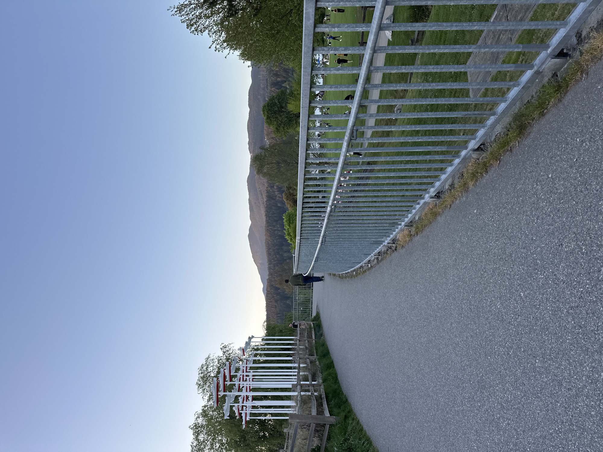 The photograph shows a slightly curved asphalt path, bordered on one side by a low metal railing and on the other by a decorative fence, next to a grass field where individuals appear to be gathered. Mountains can be seen in the background under a light blue sky. The photograph appears to have been taken in Port Moody, Canada.