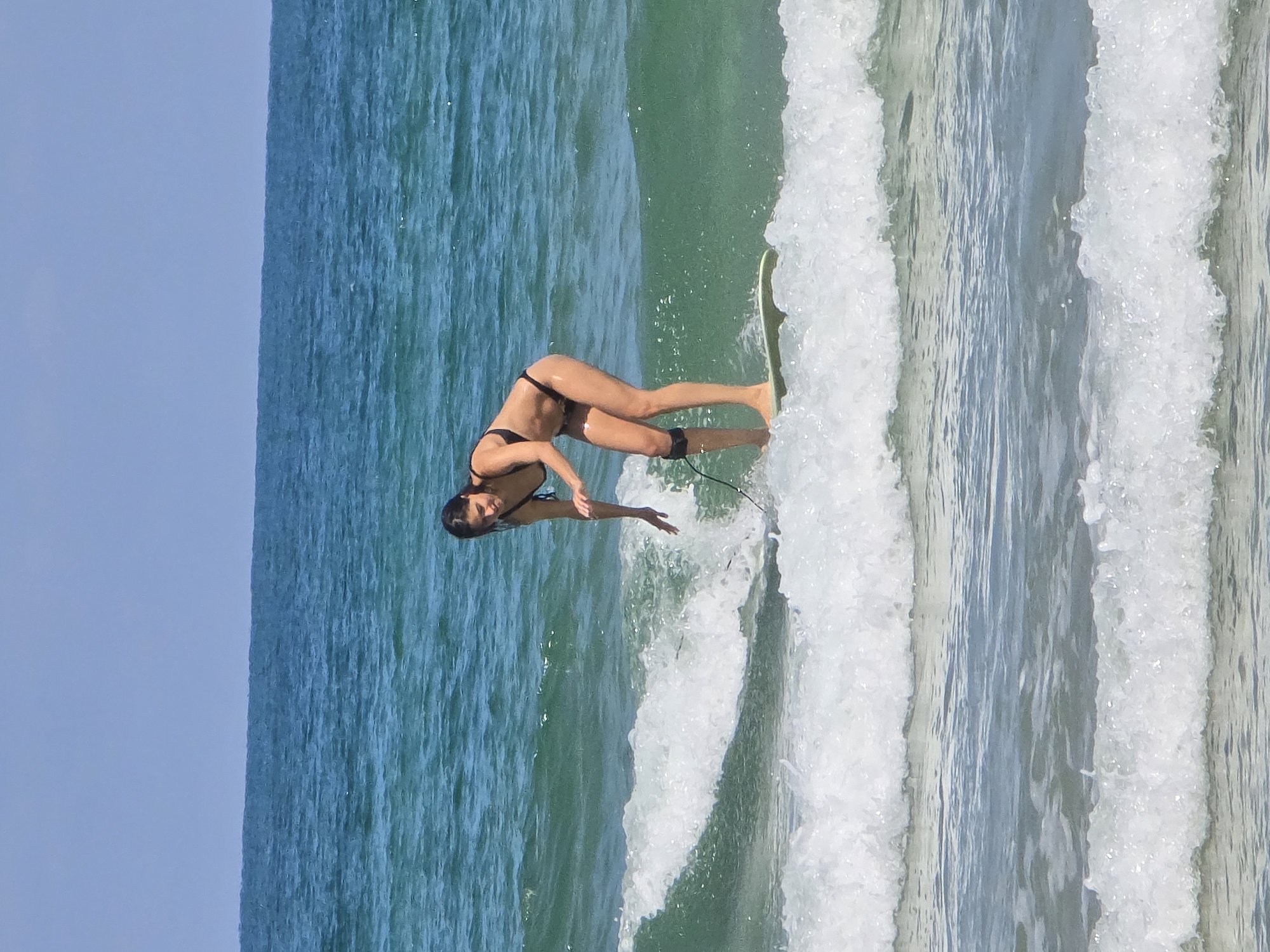 A surfer in a black bikini is lying on a surfboard in the ocean. The location appears to be near a coastline with a clear sky. The water is churning with wave action. The surfer is leaning forward, arm extended towards the water. The ocean is turquoise with white foam from the waves. It is in Tel Aviv.