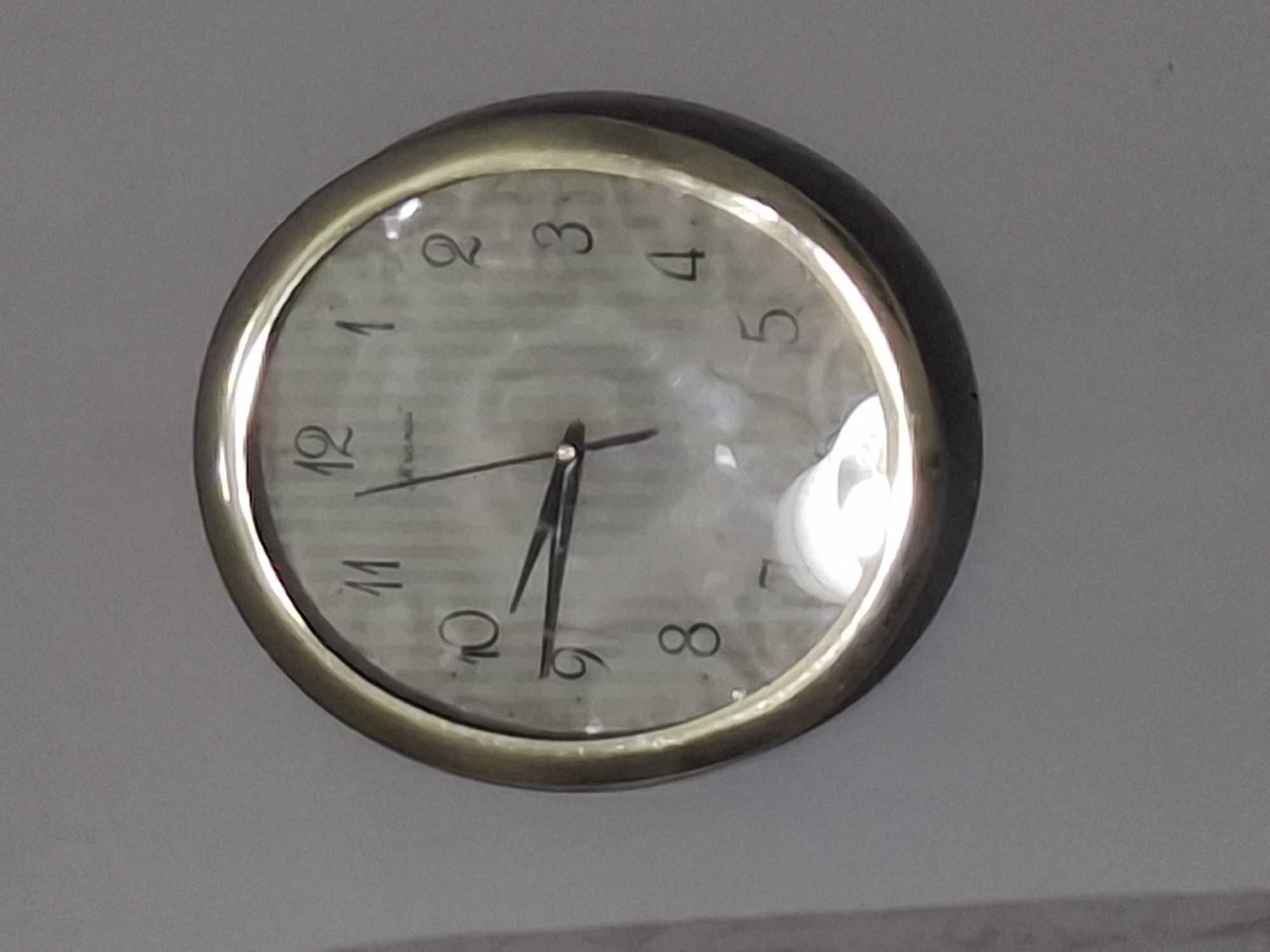 The image is a close-up, slightly angled shot of a round analog clock mounted on a plain, light gray wall. The clock has a metallic gold-colored rim and a white face with black numerals. The hands of the clock are also black. The hour hand is pointing just before the 10, and the minute hand is pointing just after the 9. The clock face has a subtle textured pattern, and there is a bright reflection on the glass cover, obscuring parts of the dial. The wall shows some texture and slight discoloration. No people or any other objects are visible in the frame. There is no discernible information or context to place this image in Almaty, Kazakhstan.
