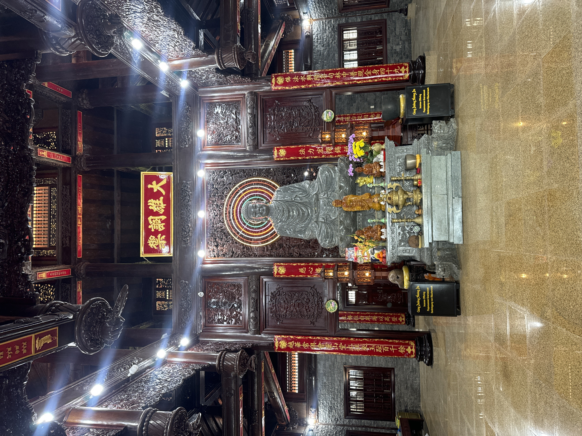 This is a full-shot, eye-level, indoor photograph of the interior of a Buddhist temple. The central focus is a large, seated Buddha statue carved from stone, positioned on a tiered altar. The Buddha is depicted with serene facial features and is adorned with a robe. Surrounding the Buddha statue are smaller statues of other deities and offerings such as flowers and incense burners.
The altar is set against a backdrop of intricately carved wooden panels, with a circular, neon-colored halo behind the Buddha. Flanking the altar are tall, dark wooden pillars with red banners bearing gold calligraphy hanging from them. The temple's architecture features elaborate dark wood carvings throughout the ceiling and walls, with red accents highlighting various decorative elements. The floor is polished and reflective, showing the illumination from multiple light fixtures strategically placed throughout the space. To the left and right of the main altar, there are stone walls with recessed windows. In the foreground, two black boxes with yellow text are placed on the floor. The overall impression is one of grandeur and solemnity. This image is likely taken at the Quán Âm Tu Viện temple in Thuận Nam, Ninh Thuận province, Vietnam.