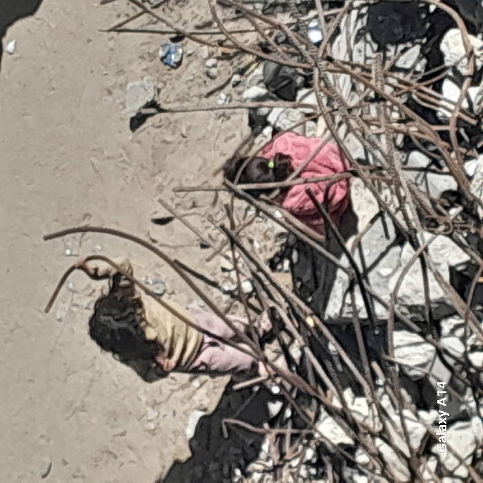 The image displays an outdoor scene in Khan Yunis, Palestinian Territory, characterized by extensive demolition debris. The ground consists of large and small fragments of concrete, stones, and loose soil or dust. Numerous rusted, bent, and broken steel rebar rods are scattered across the scene, particularly prominent in the foreground and midground, some standing upright.
Two individuals, consistent with children, are present. On the left, a child stands with their back to the camera, possessing long, dark, wavy hair. They wear a light-colored, long-sleeved top and light pink or purple trousers. Their left arm is bent at the elbow. To the right, a second child is seated on the ground, partially obscured by rebar. This child is wearing a pink garment, and dark hair is visible. A small, bright green object is visible near their face or hand. Harsh shadows are cast across the rubble, indicating strong overhead lighting. Sparse debris, including a crumpled item, is visible in the background. A "Galaxy A14" watermark is present in the bottom-left corner of the image.