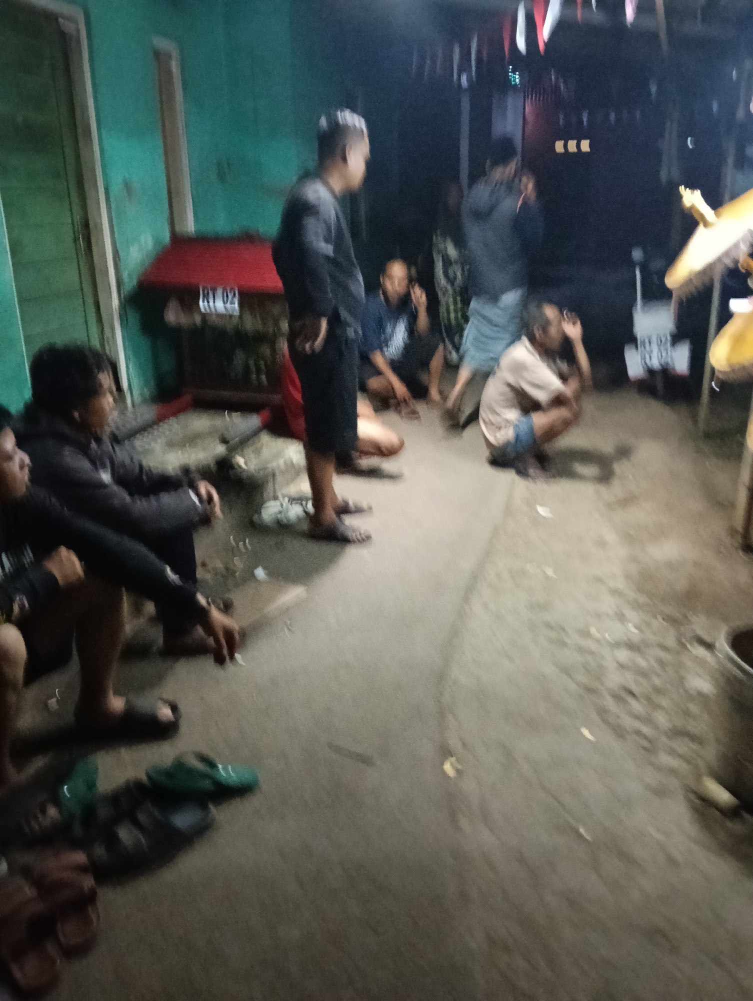 In an outdoor, dimly lit environment with a concrete ground, several Indonesian individuals are gathered. The scene suggests a casual social gathering, possibly in front of a residence, given the presence of a green wall and a red structure with "RT 02" visible. Most individuals are seated or crouching, some are smoking, and their clothing appears to be everyday attire. A yellow umbrella-like object is partially visible on the right, and some footwear, including sandals, are scattered on the ground. The overall impression is of a relaxed, informal gathering in a residential area, likely in Tasikmalaya, Indonesia.
